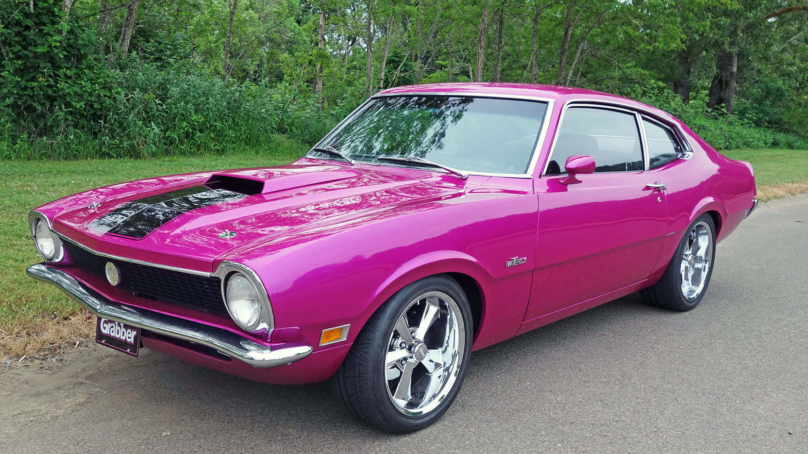 Ford Maverick 桌布 壁紙 — 一輛充滿活力的粉紅色經典肌肉車，配備鍍鉻輪圈，停在鋪砌的道路上，旁邊是茂密的綠樹。