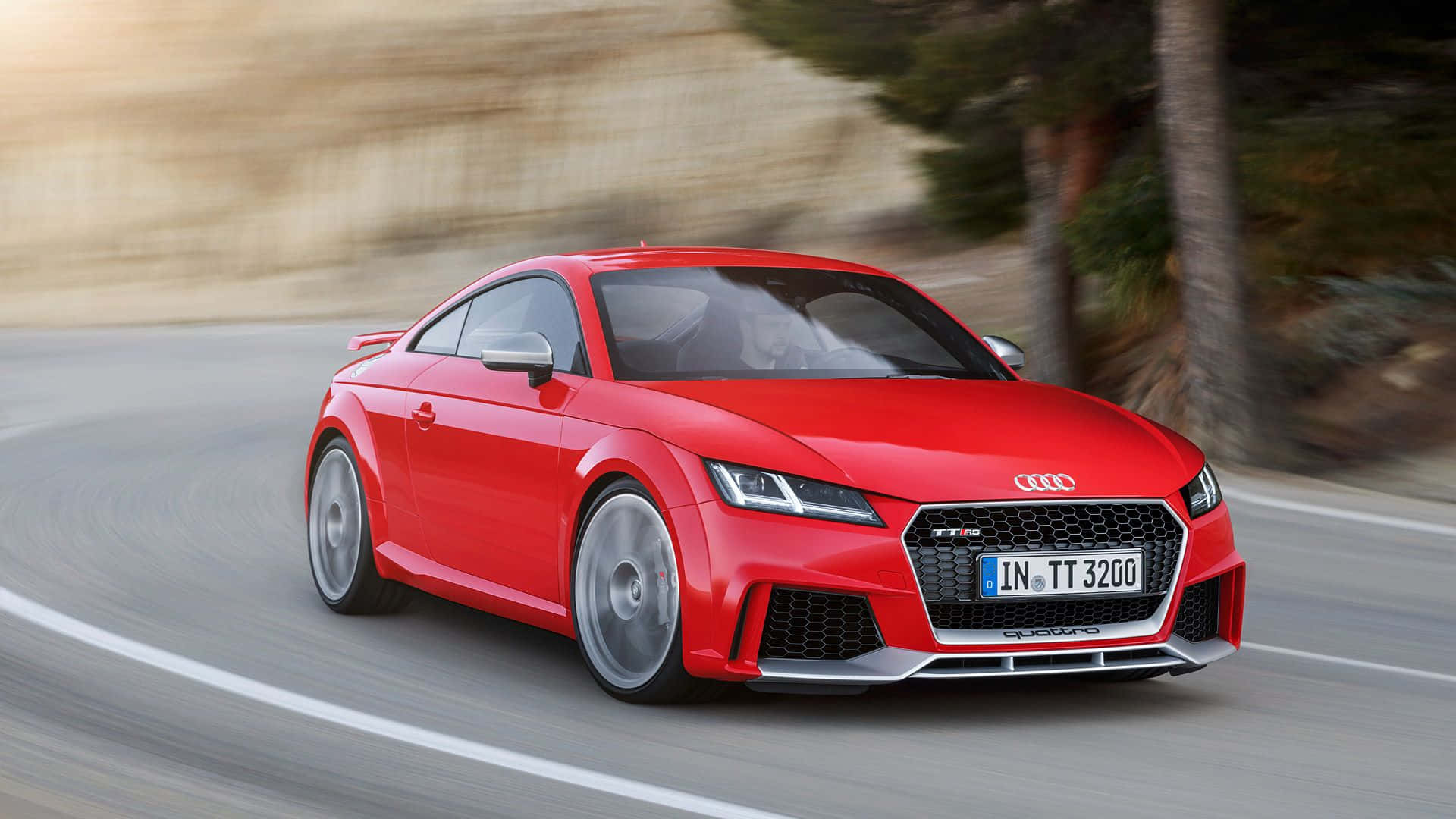 Audi Tt 桌布 壁紙 — 一輛充滿活力的紅色 Audi TT RS 在蜿蜒的道路上疾馳，背景是模糊的樹木和山丘。