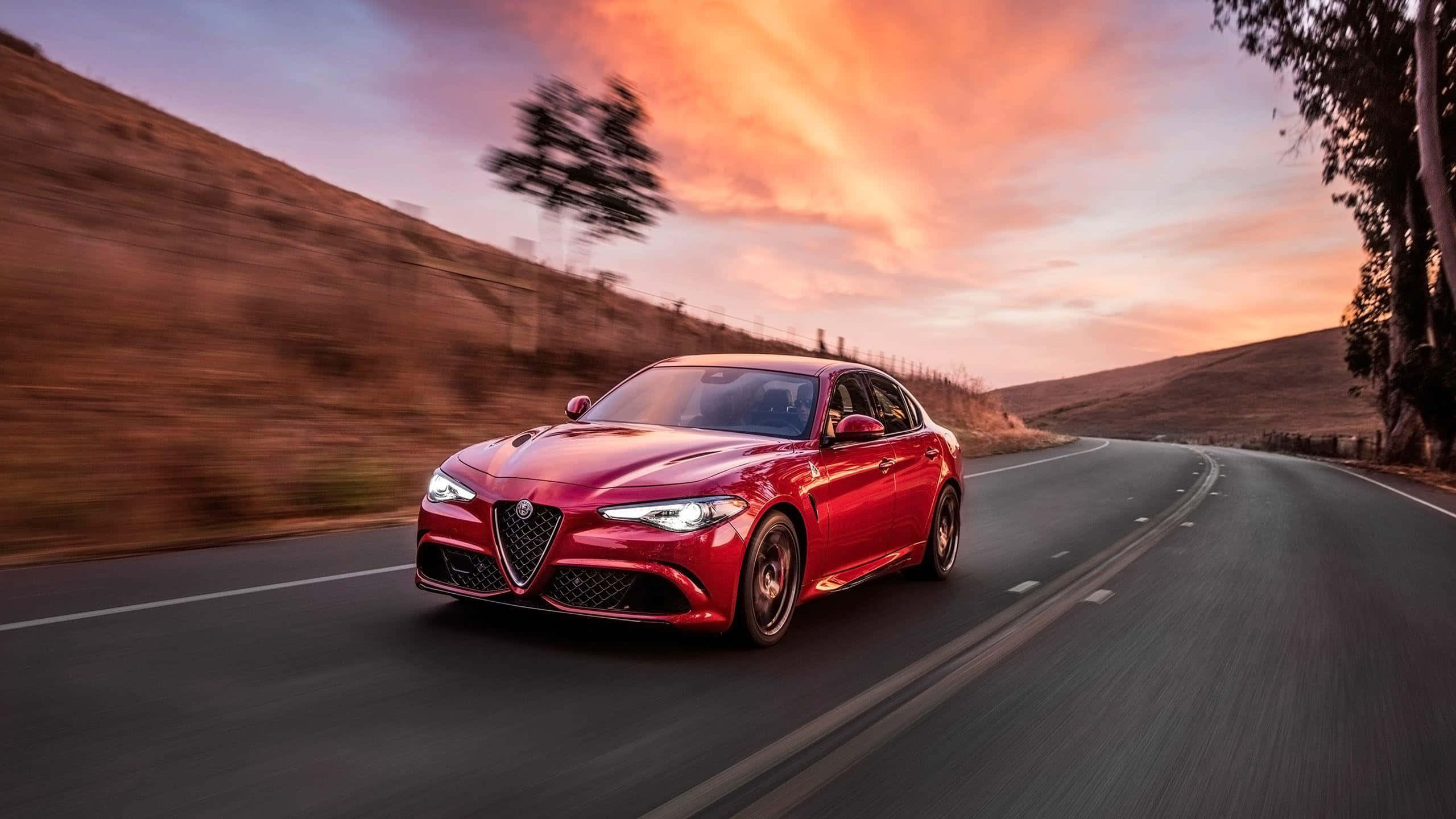 Alfa Romeo Giulia 桌布 壁紙 — 一輛充滿活力的紅色Alfa Romeo房車在火紅的日落天空下，沿著蜿蜒的道路疾馳，兩旁的山丘模糊不清。