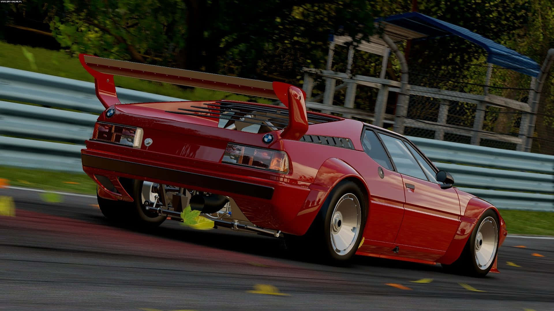 電玩遊戲 桌布 壁紙 — 一輛充滿活力的紅色BMW M1賽車在賽道上疾馳，背景有動態模糊和綠色樹葉。