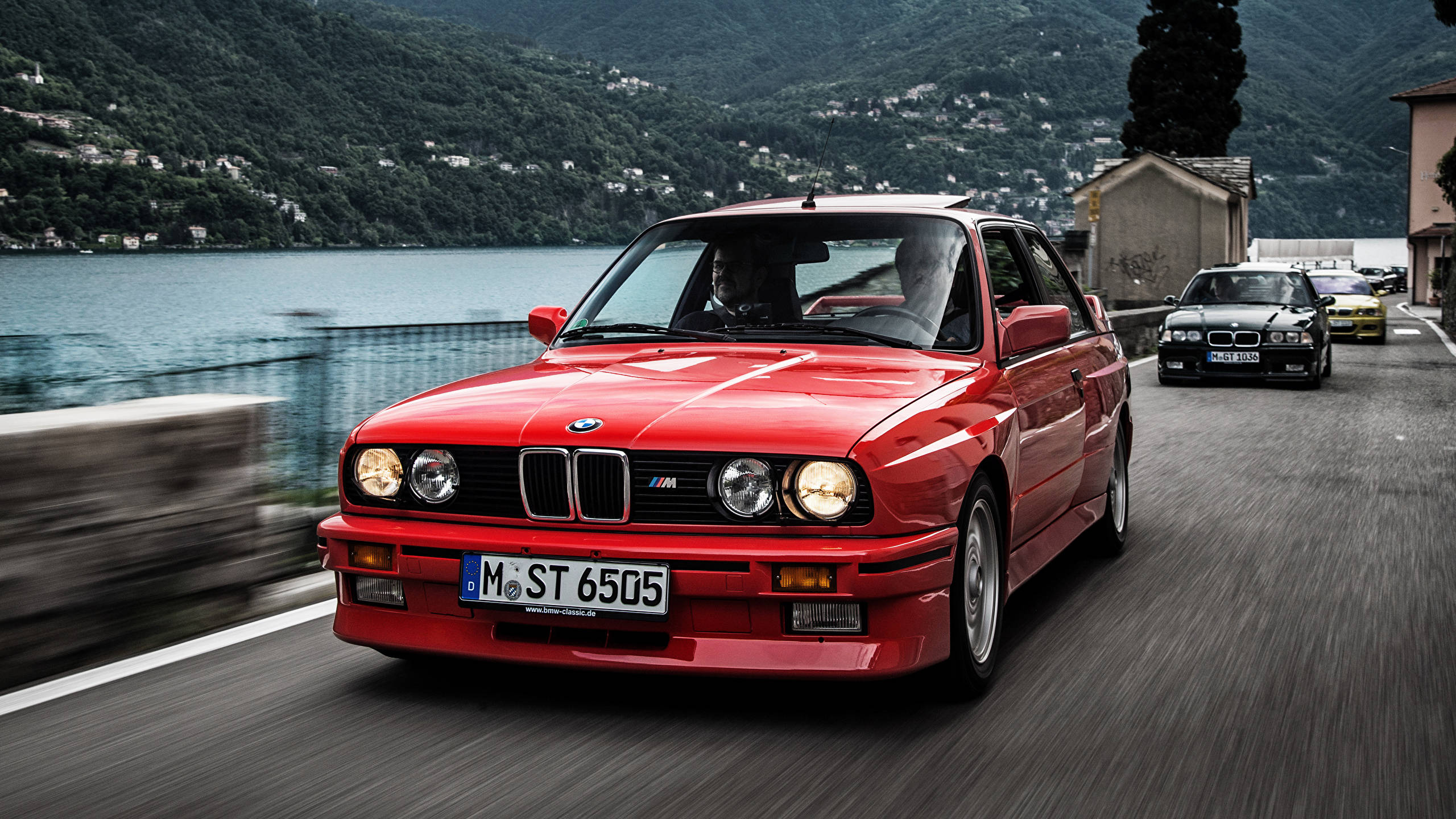 classic bmw 桌布 壁紙 — 一輛充滿活力的紅色BMW M3沿著湖邊道路疾馳，帶有動態模糊效果，兩旁有其他車輛和茂密的綠色山丘。