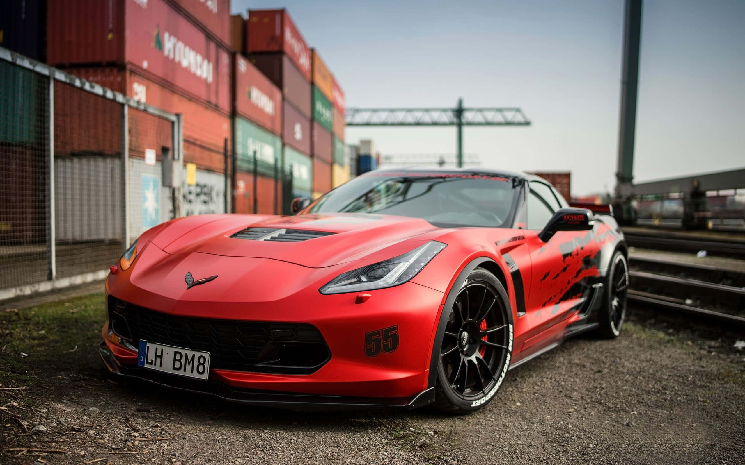 Chevrolet Corvette Z06 桌布 壁紙 — 一輛充滿活力的紅色Corvette跑車，帶有黑色裝飾和55號碼，停在碎石上，靠近貨櫃和鐵路軌道。