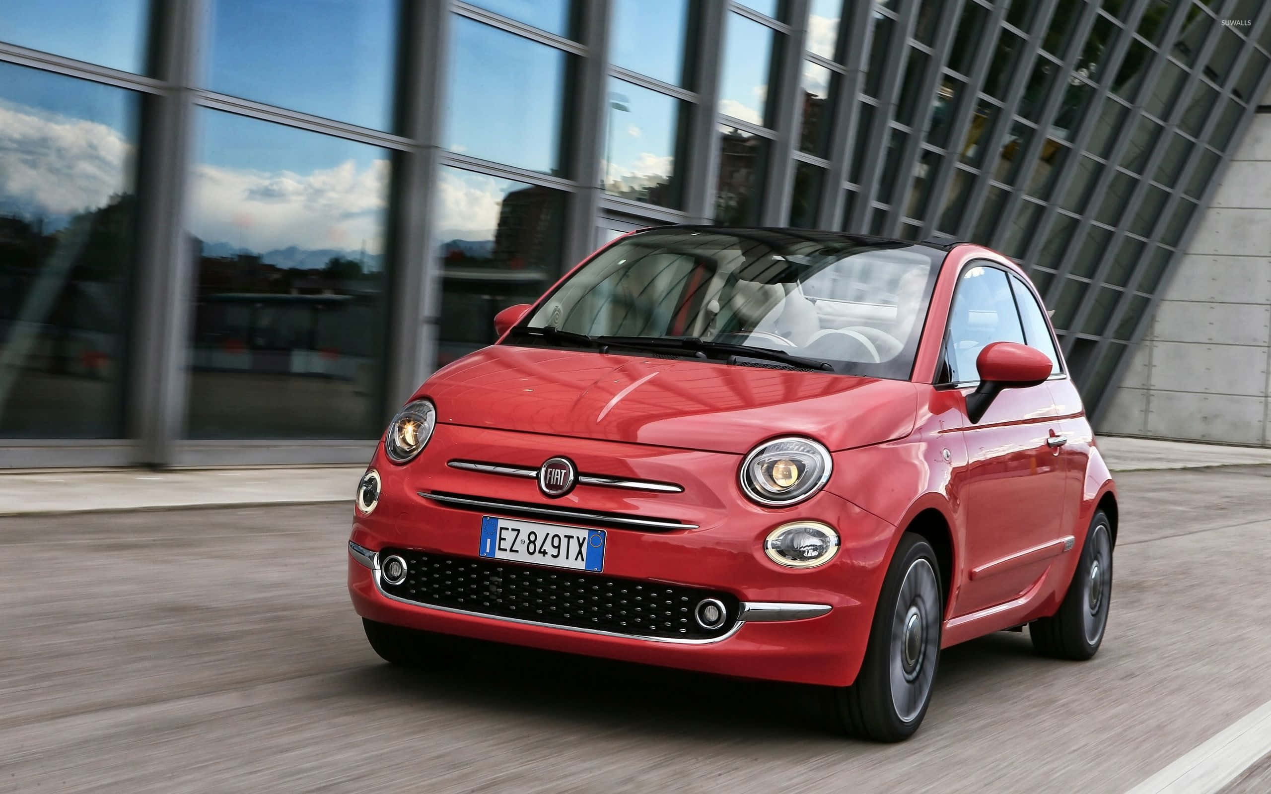 Fiat 500l 桌布 壁紙 — 一輛充滿活力的紅色Fiat 500駛過現代玻璃建築，路上帶有動態模糊效果。