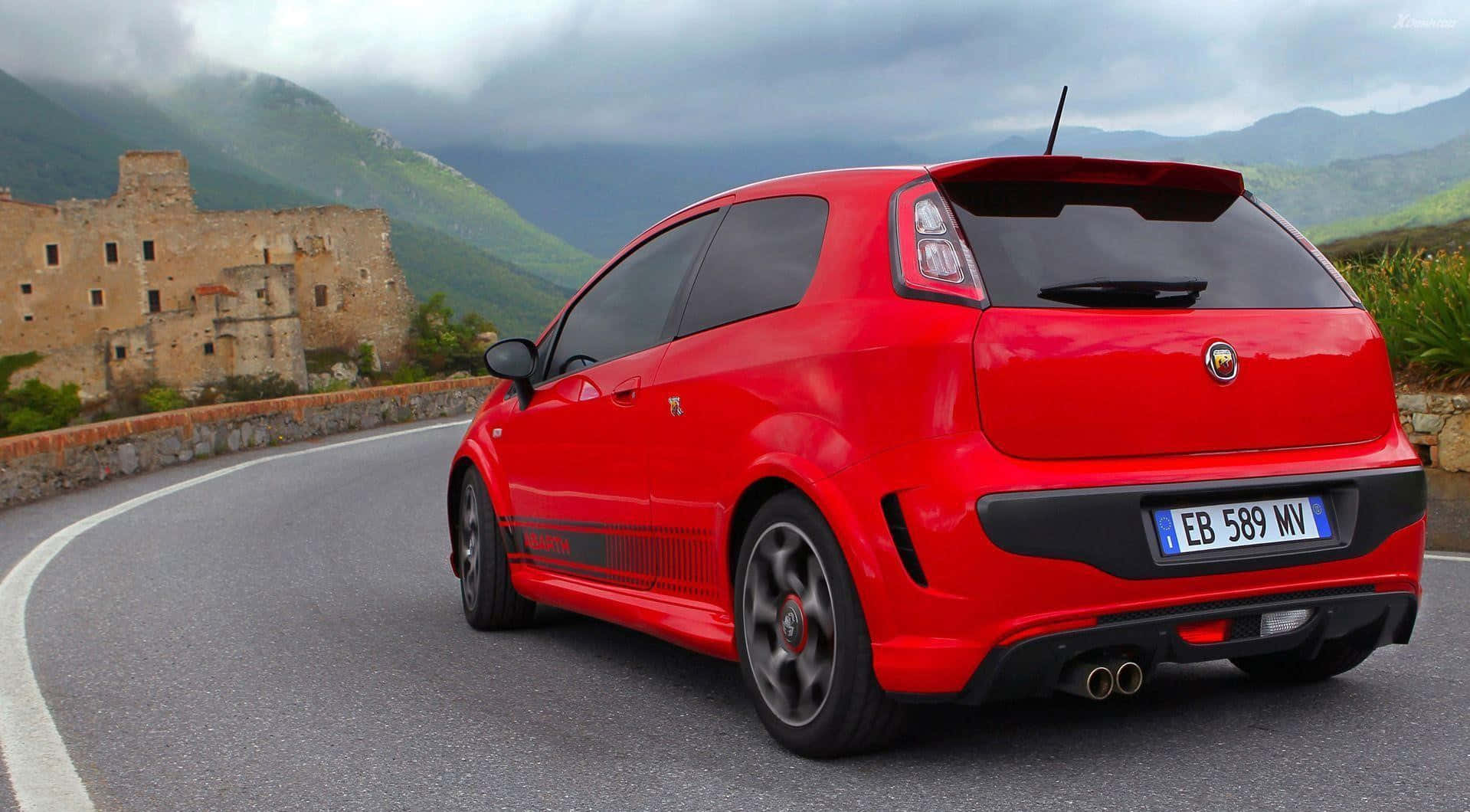Abarth 桌布 壁紙 — 一輛充滿活力的紅色Fiat Punto Evo ABARTH在陰天沿著蜿蜒的山路飛馳，經過一座古老的石頭廢墟。