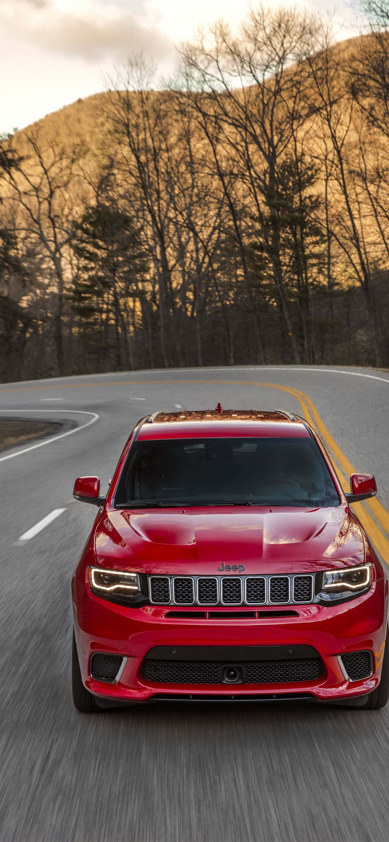 Jeep Trackhawk 桌布 壁紙 — 一輛充滿活力的紅色Jeep Grand Cherokee Trackhawk在黃金時段沿著蜿蜒的山路疾馳，背景是光禿禿的樹木和山丘。