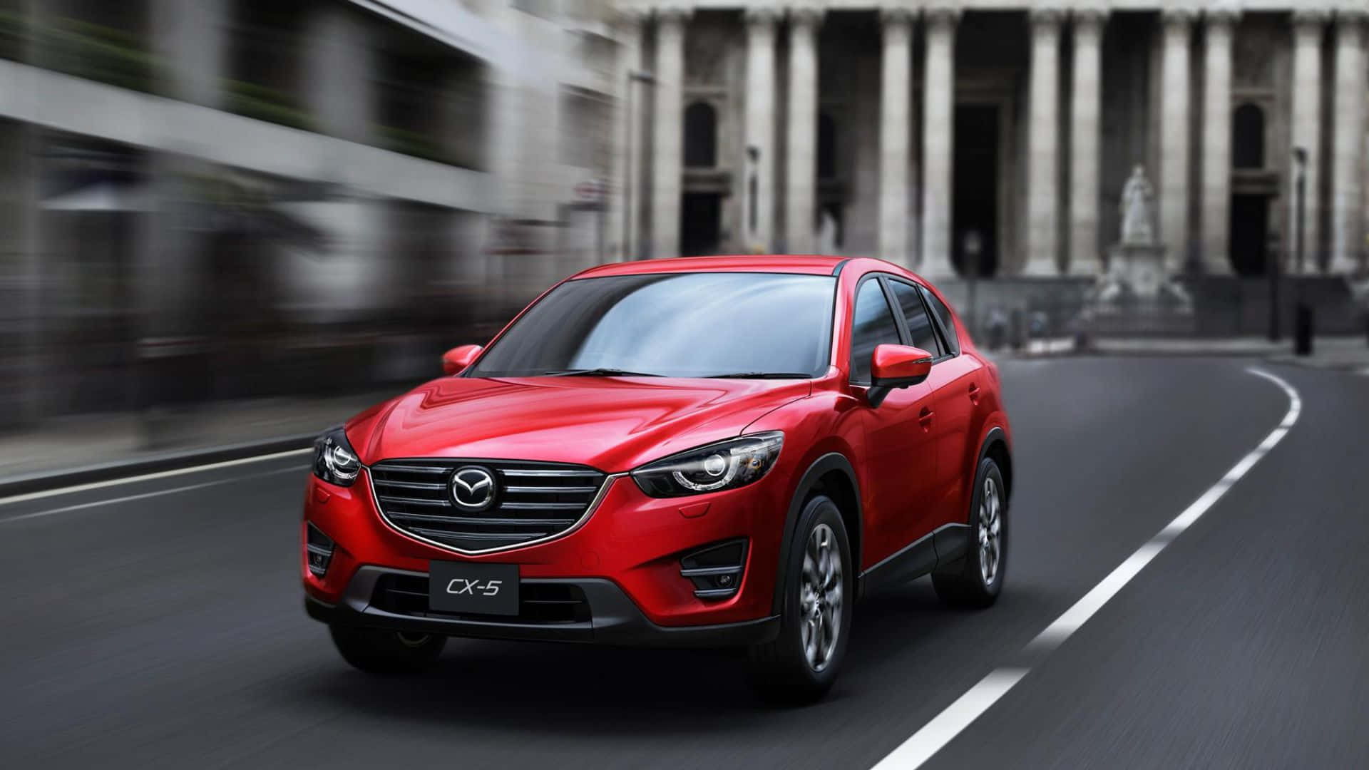 Mazda Cx-5 桌布 壁紙 — 一輛充滿活力的紅色Mazda CX-5 SUV在帶有動態模糊背景和古典建築的城市街道上飛馳。
