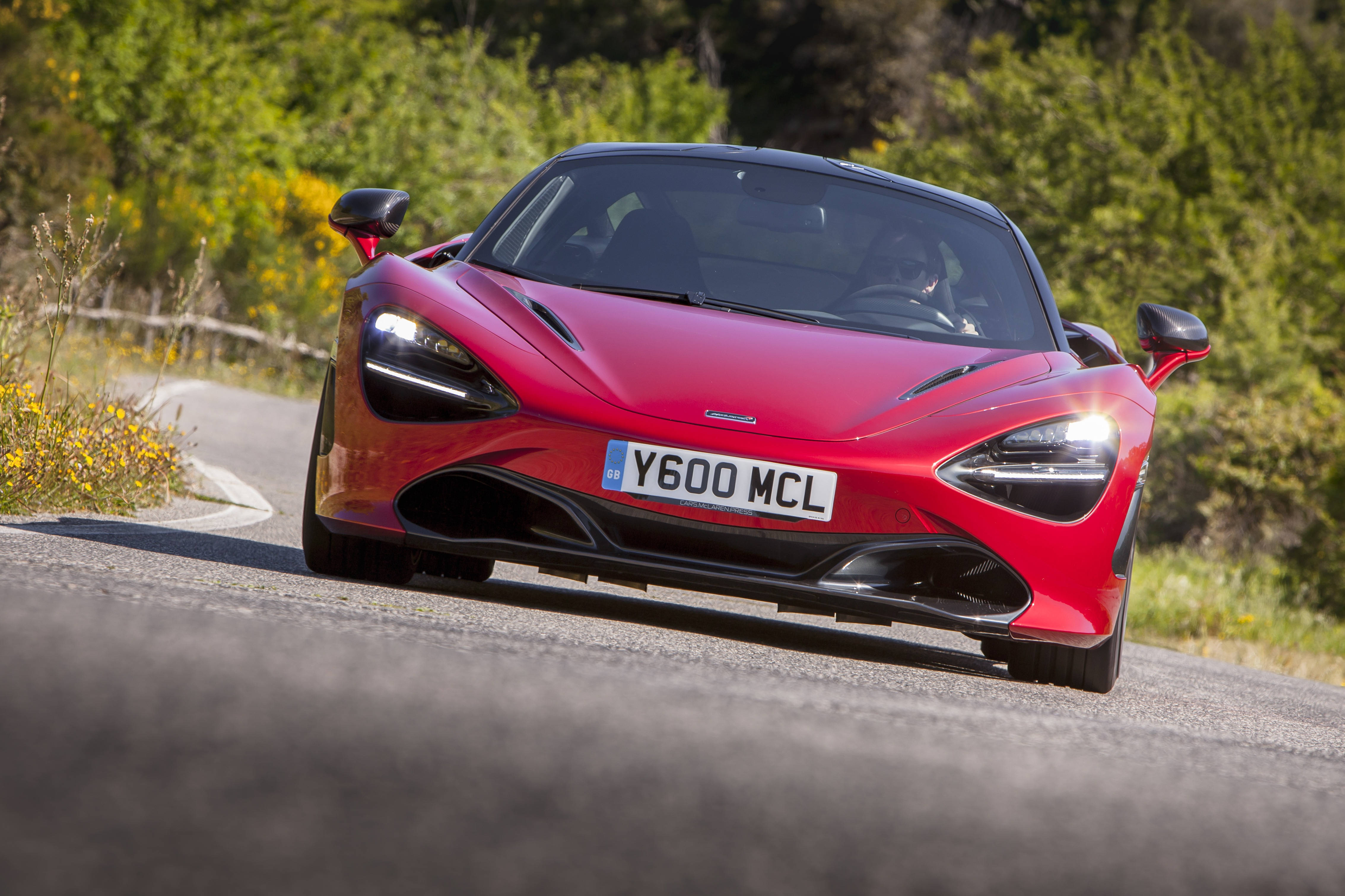 mclaren 720s 4K 桌布 壁紙 — 一輛充滿活力的紅色McLaren 720S跑車在明媚陽光下，沿著一條蜿蜒的鄉村道路飛馳，道路兩旁綠樹成蔭，黃色野花盛開。