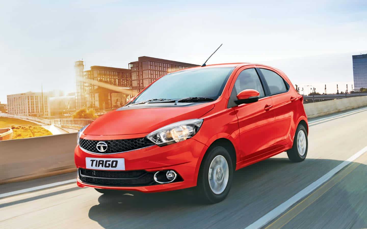 Tata Motors 桌布 壁紙 — 一輛充滿活力的紅色Tata Tiago掀背車在陽光明媚的城市高速公路上奔馳，背景是現代建築。