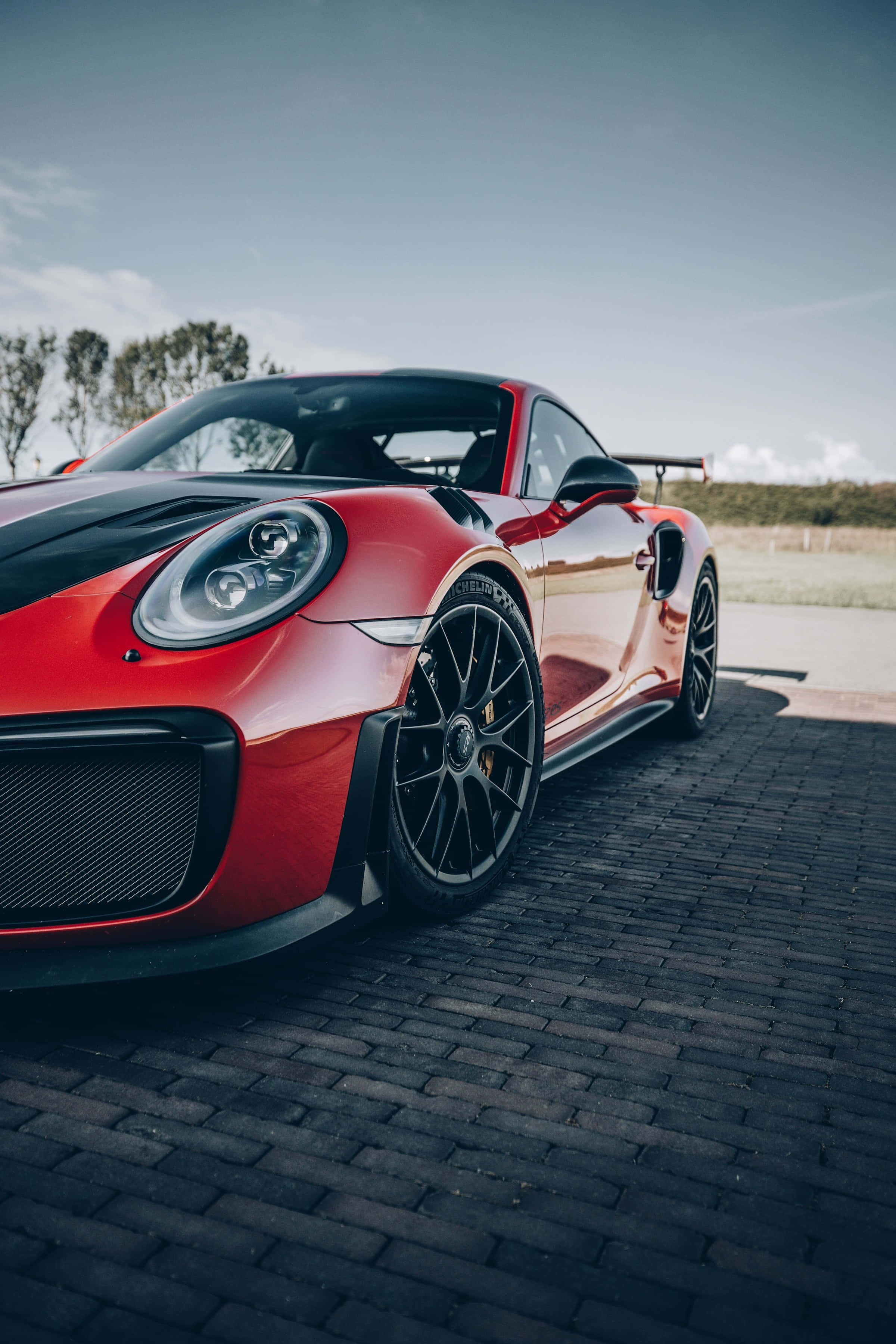 cars 桌布 壁紙 — 一輛充滿活力的紅色保時捷911 GT2 RS，帶有黑色點綴，停泊在晴朗天空下的鵝卵石路面上。