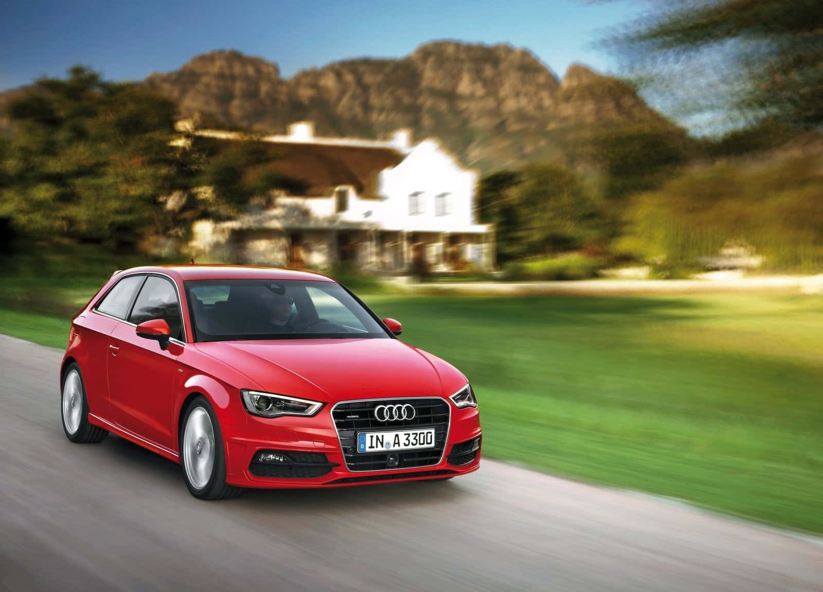 Audi A3 桌布 壁紙 — 一輛充滿活力的紅色奧迪A3沿著蜿蜒的道路飛馳，經過一棟白色房子，背景是山脈。