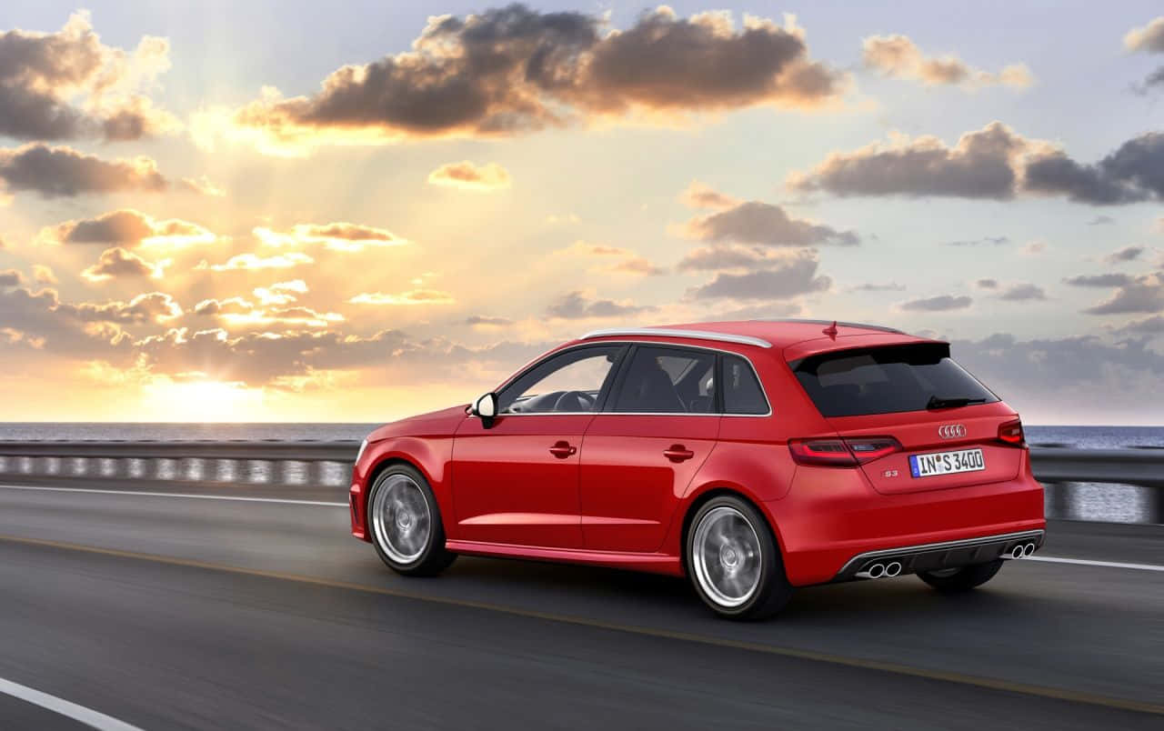 Audi S3 桌布 壁紙 — 一輛充滿活力的紅色奧迪旅行車沿著海岸公路行駛，在壯觀的日落天空下，金色雲彩點綴其間。