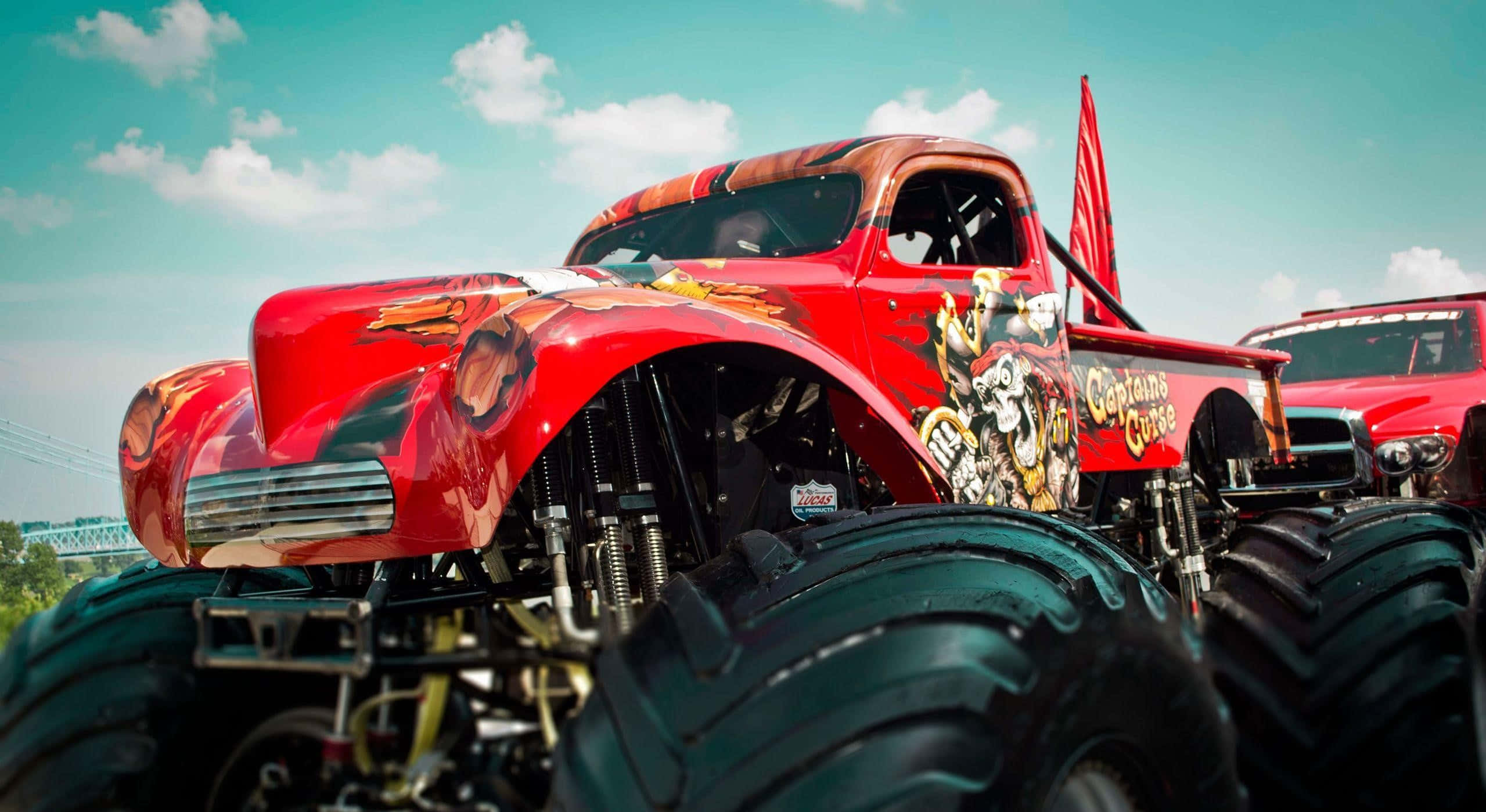 Cool Car 桌布 壁紙 — 一輛充滿活力的紅色怪獸卡車，配備巨大的黑色輪胎和骷髏圖案，在藍天白雲下的陽光戶外場景中佔據主導地位。