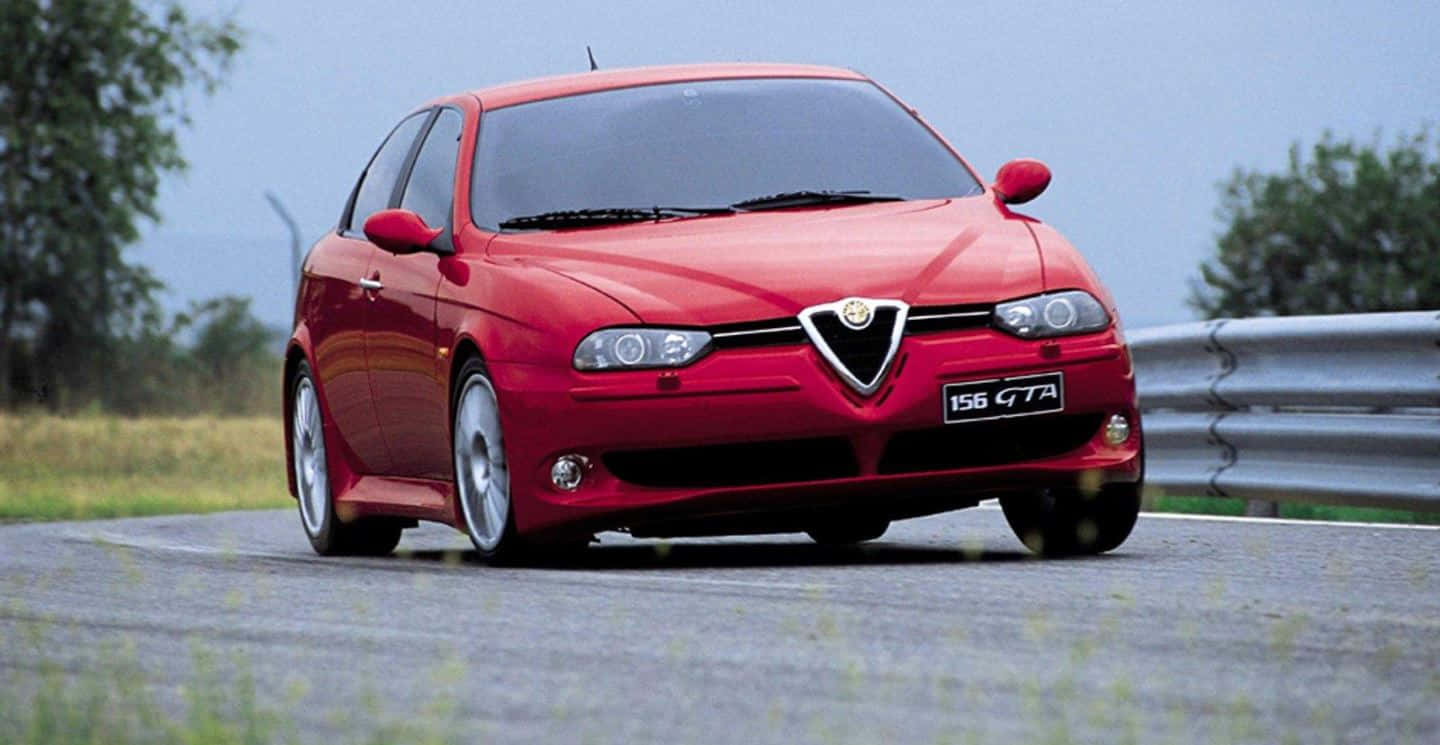 Alfa Romeo 156 桌布 壁紙 — 一輛充滿活力的紅色愛快羅密歐156 GTA在陰天之下，沿著一條彎曲的瀝青道路飛馳，道路兩旁有護欄和綠樹。