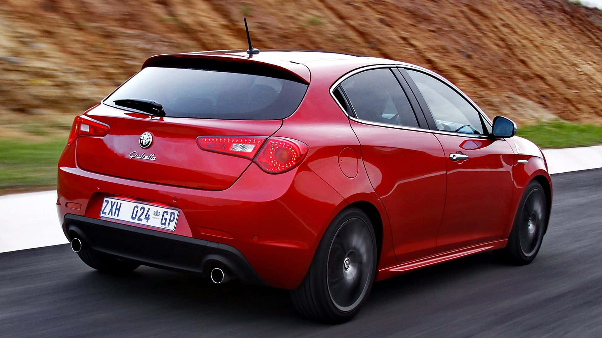 Alfa Romeo Giulietta 桌布 壁紙 — 一輛充滿活力的紅色愛快羅密歐Giulietta掀背車在蜿蜒的道路上行駛，背景是模糊的土色山丘。