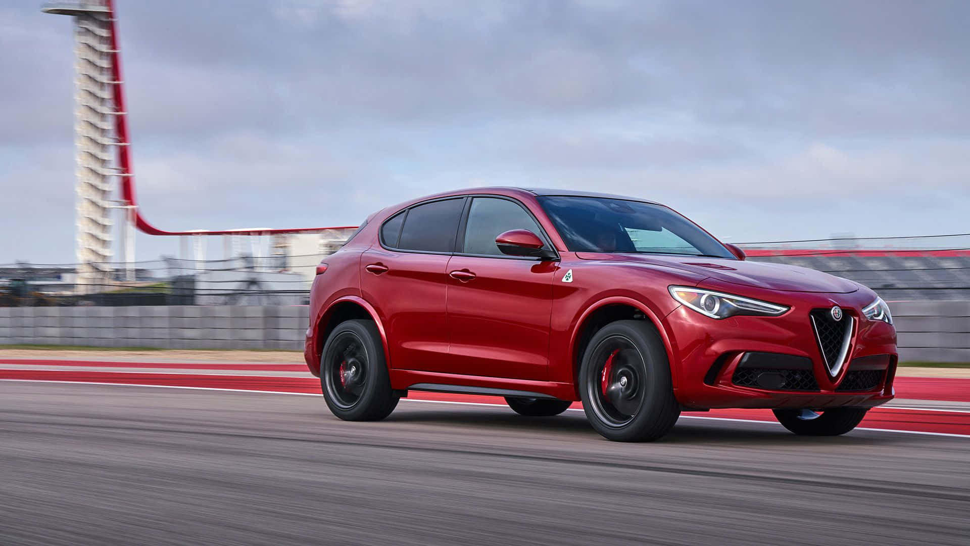 Alfa Romeo Stelvio 桌布 壁紙 — 一輛充滿活力的紅色愛快羅密歐SUV在賽道上高速行駛，帶有動態模糊效果，背景有一座高塔。