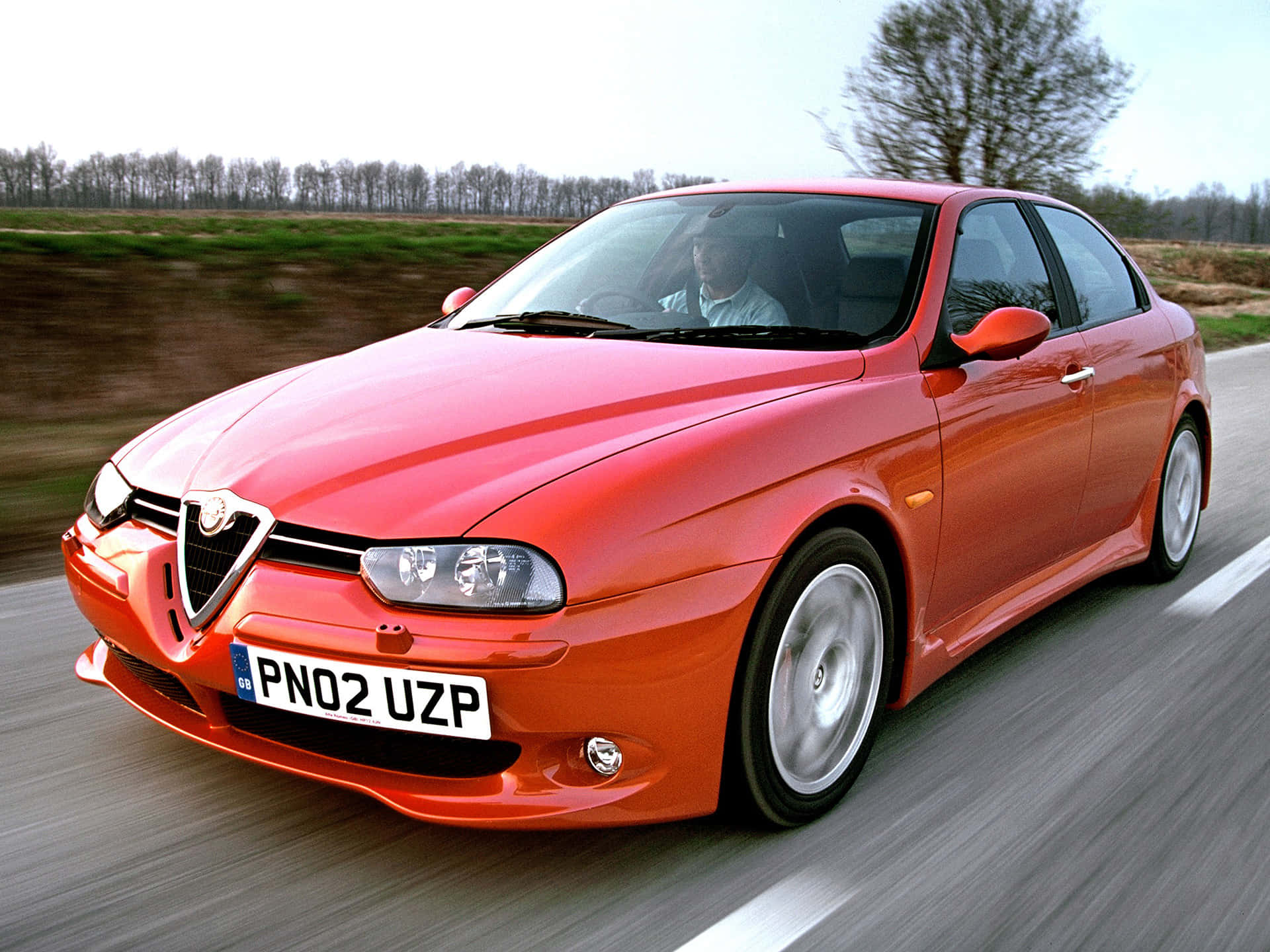 Alfa Romeo 156 桌布 壁紙 — 一輛充滿活力的紅色愛快羅密歐跑車在鄉村道路上疾馳，帶有模糊的動態效果，車內駕駛員清晰可見。