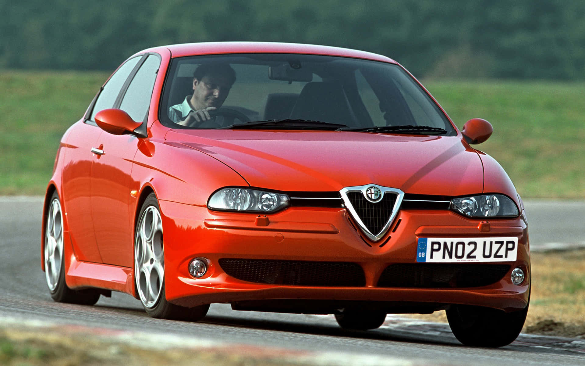 Alfa Romeo 156 桌布 壁紙 — 一輛充滿活力的紅色愛快羅密歐跑車，在蜿蜒的道路上疾馳，司機專注地握著方向盤。