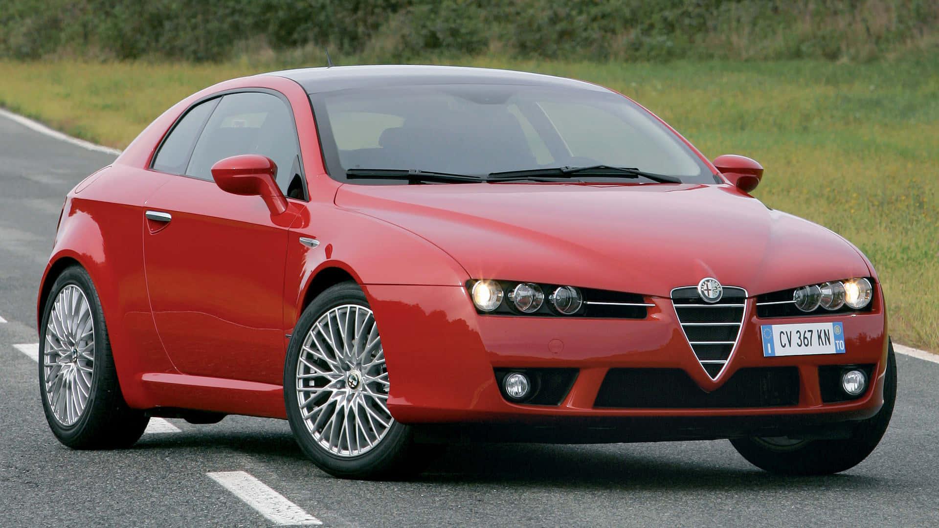 Alfa Romeo Brera 桌布 壁紙 — 一輛充滿活力的紅色愛快羅密歐跑車沿著一條蜿蜒的瀝青道路行駛，兩旁是茂密的綠草。