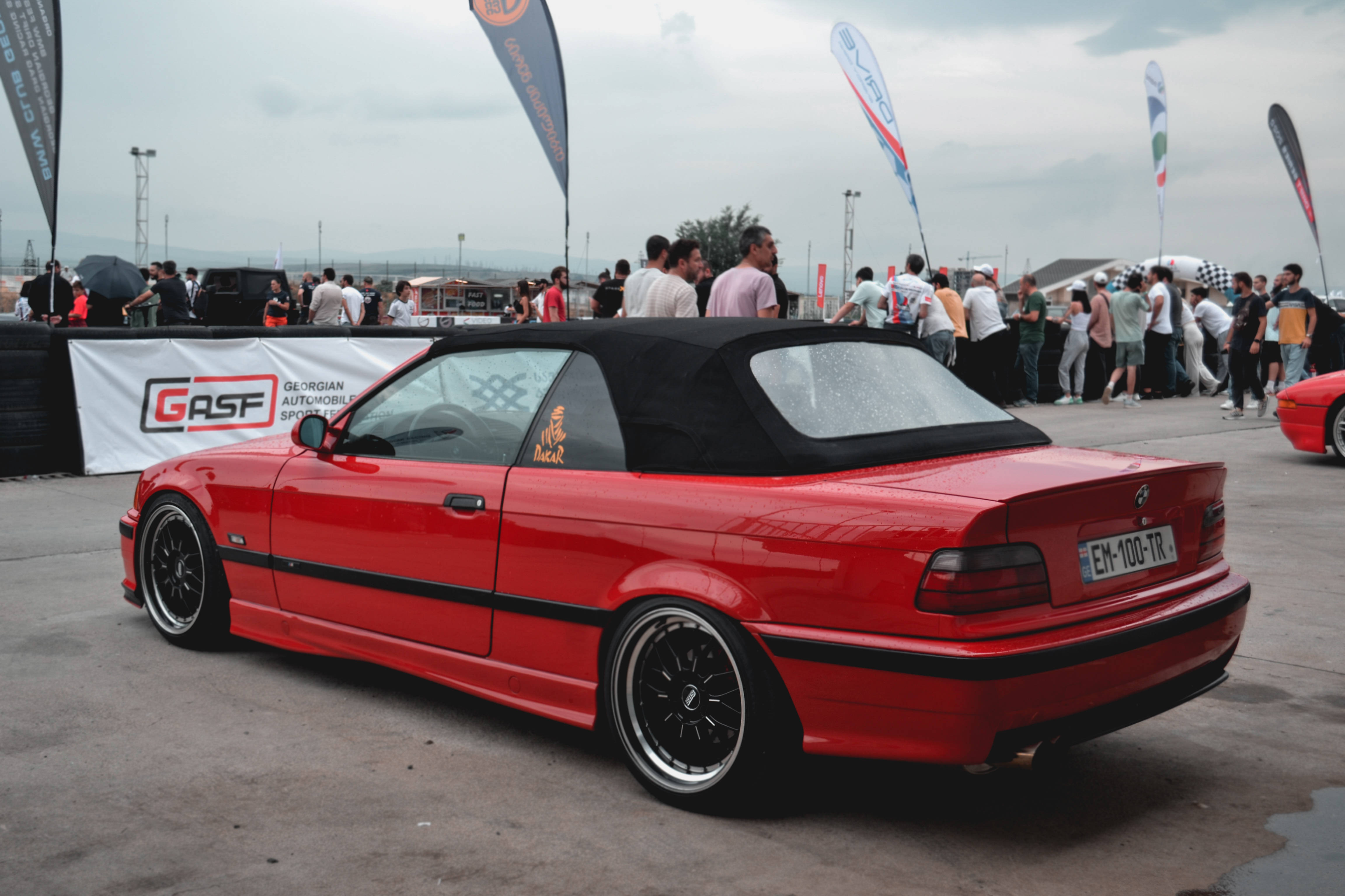 Racing 桌布 壁紙 — 一輛充滿活力的紅色敞篷跑車，帶有黑色飾邊和定制輪圈，停放在戶外汽車活動中，背景有觀眾。