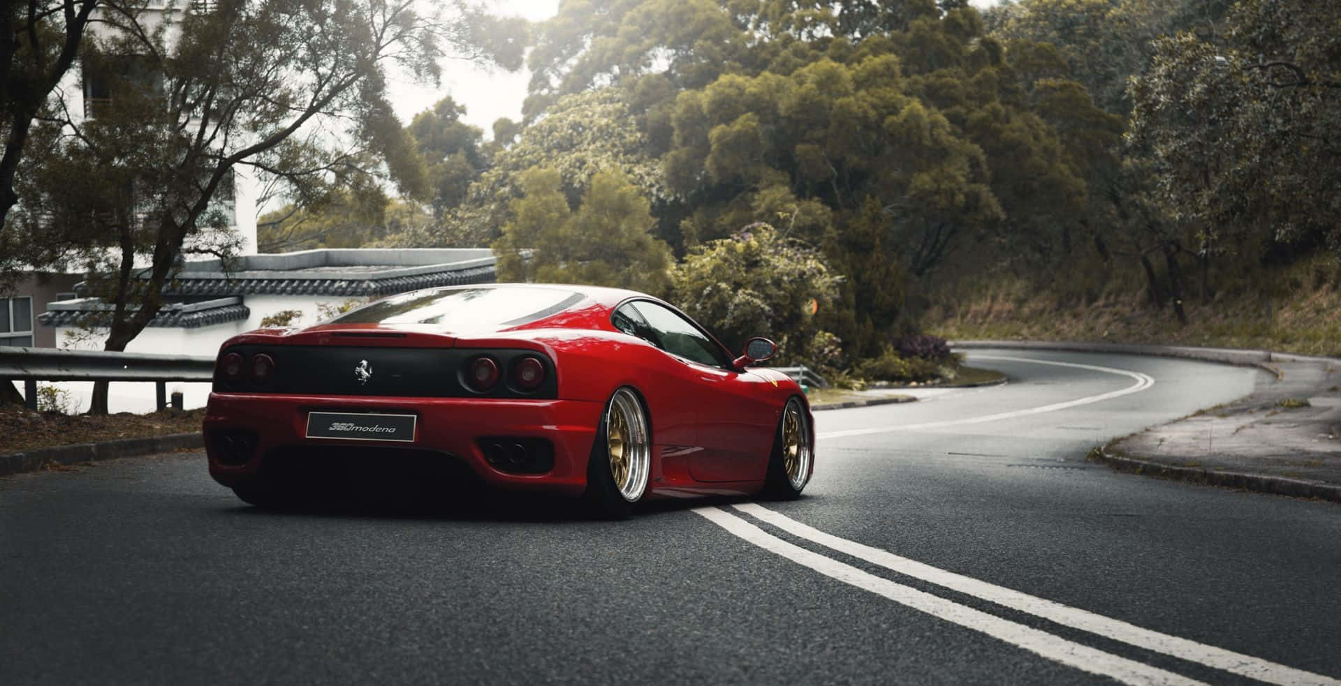 Ferrari 360 Modena 桌布 壁紙 — 一輛充滿活力的紅色法拉利F430跑車，配備金色輪圈，沿著一條蜿蜒的瀝青道路行駛，周圍環繞著茂密的綠色樹木。