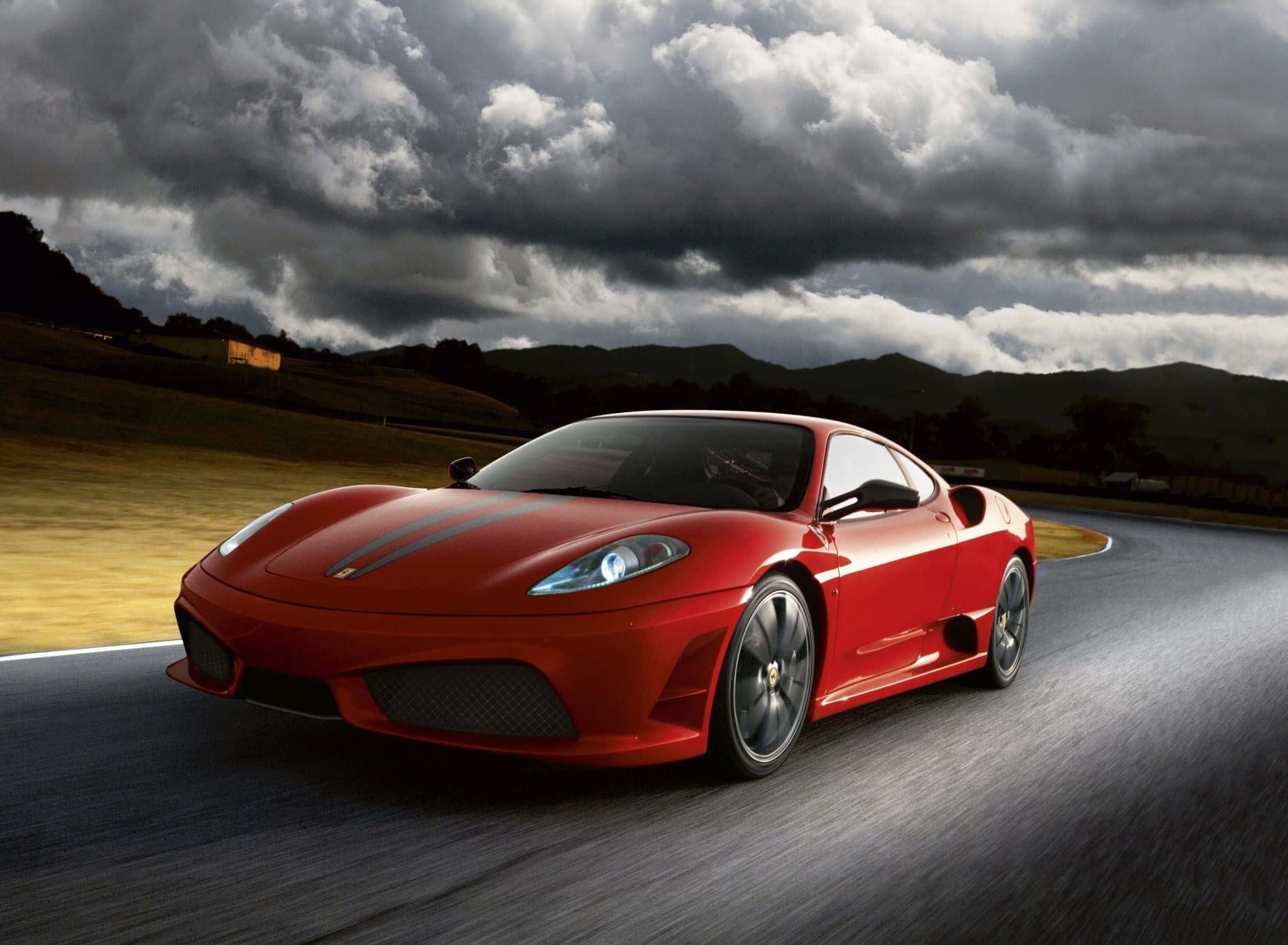 Ferrari F430 桌布 壁紙 — 一輛充滿活力的紅色法拉利跑車在戲劇性的多雲天空下沿著蜿蜒的道路疾馳，背景是起伏的丘陵。
