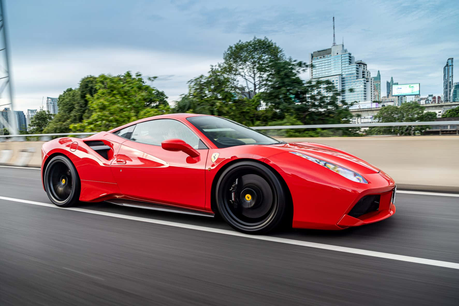 Ferrari 488 Spider 桌布 壁紙 — 一輛充滿活力的紅色法拉利跑車在現代摩天大樓和綠樹成蔭的城市高速公路上飛馳。