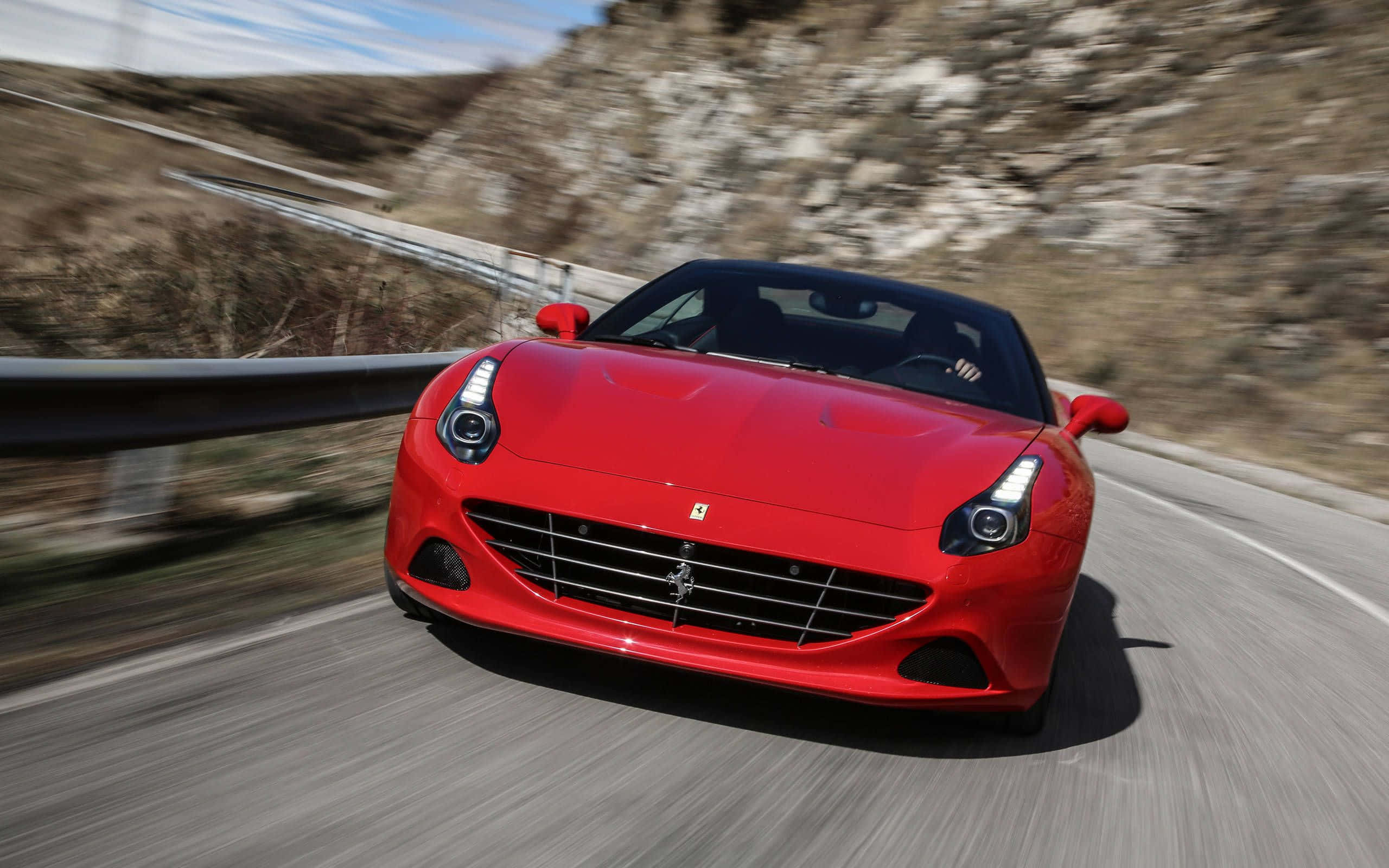 Ferrari California T 桌布 壁紙 — 一輛充滿活力的紅色法拉利跑車在蜿蜒的山路上疾馳，帶有模糊的動感和岩石地形。