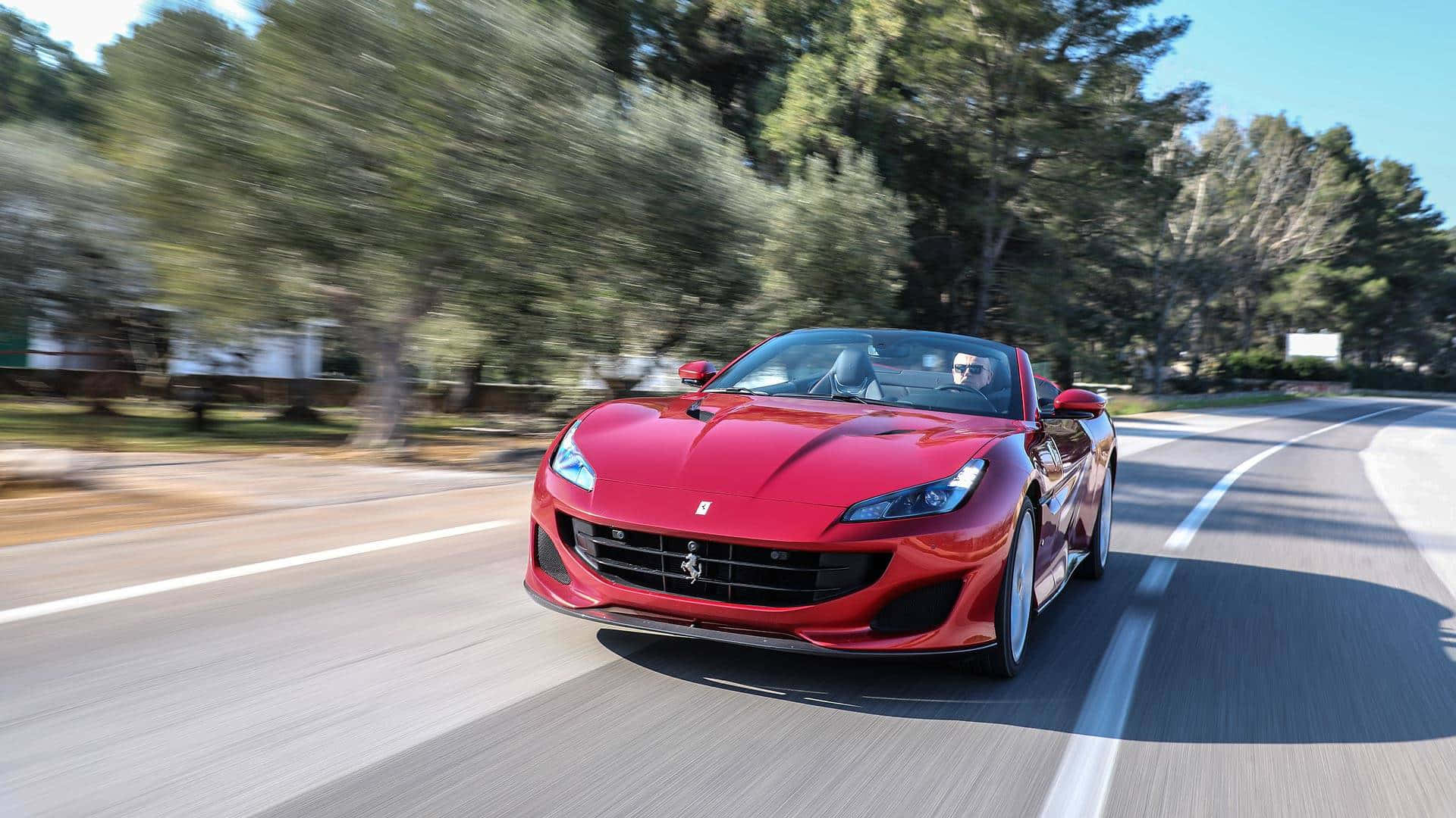 Ferrari Portofino 桌布 壁紙 — 一輛充滿活力的紅色法拉利跑車在陽光明媚的蜿蜒道路上飛馳，周圍環繞著茂密的綠樹。