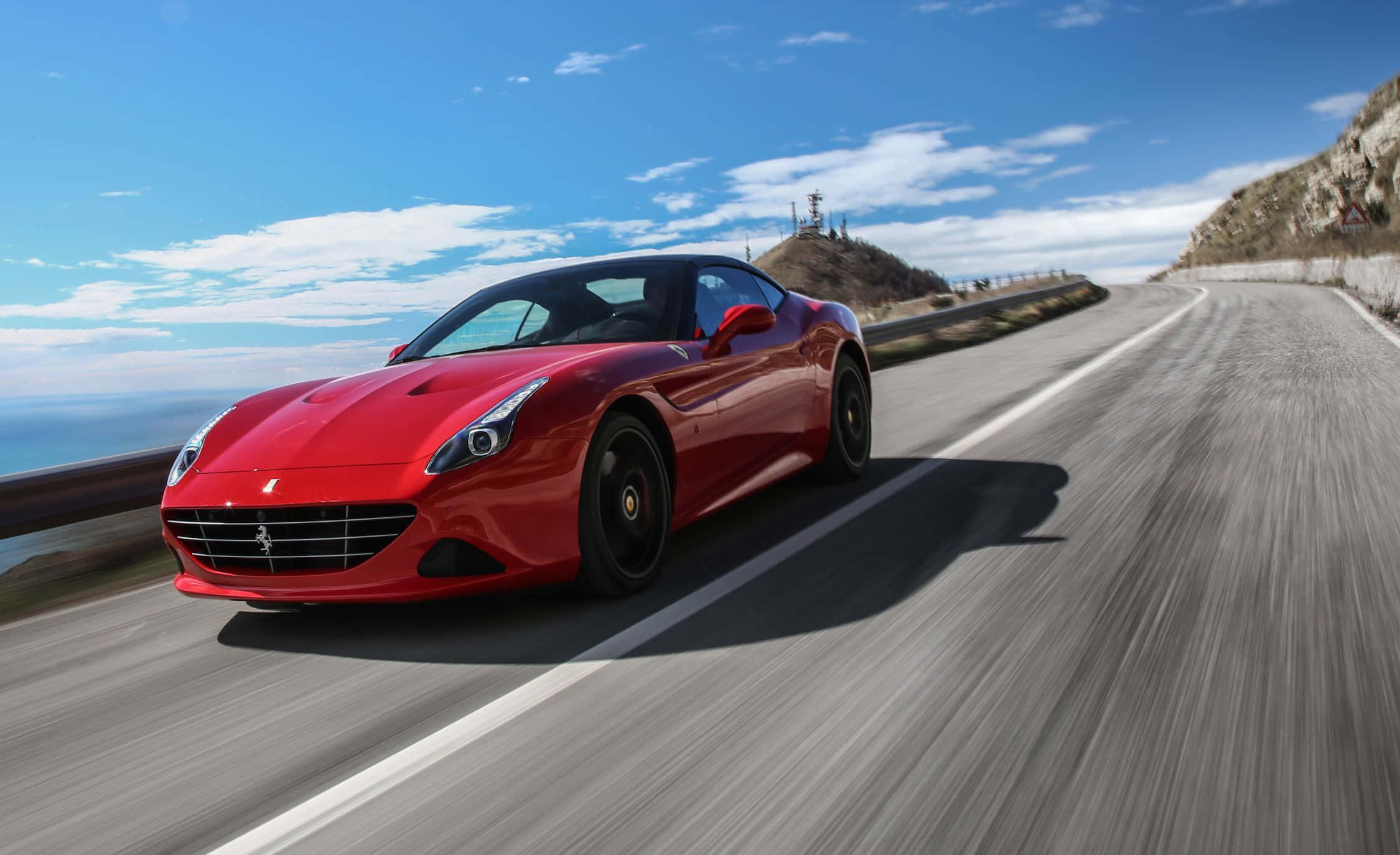 Ferrari California T 桌布 壁紙 — 一輛充滿活力的紅色法拉利跑車沿著沿海山路疾馳，天空湛藍，雲朵點綴。