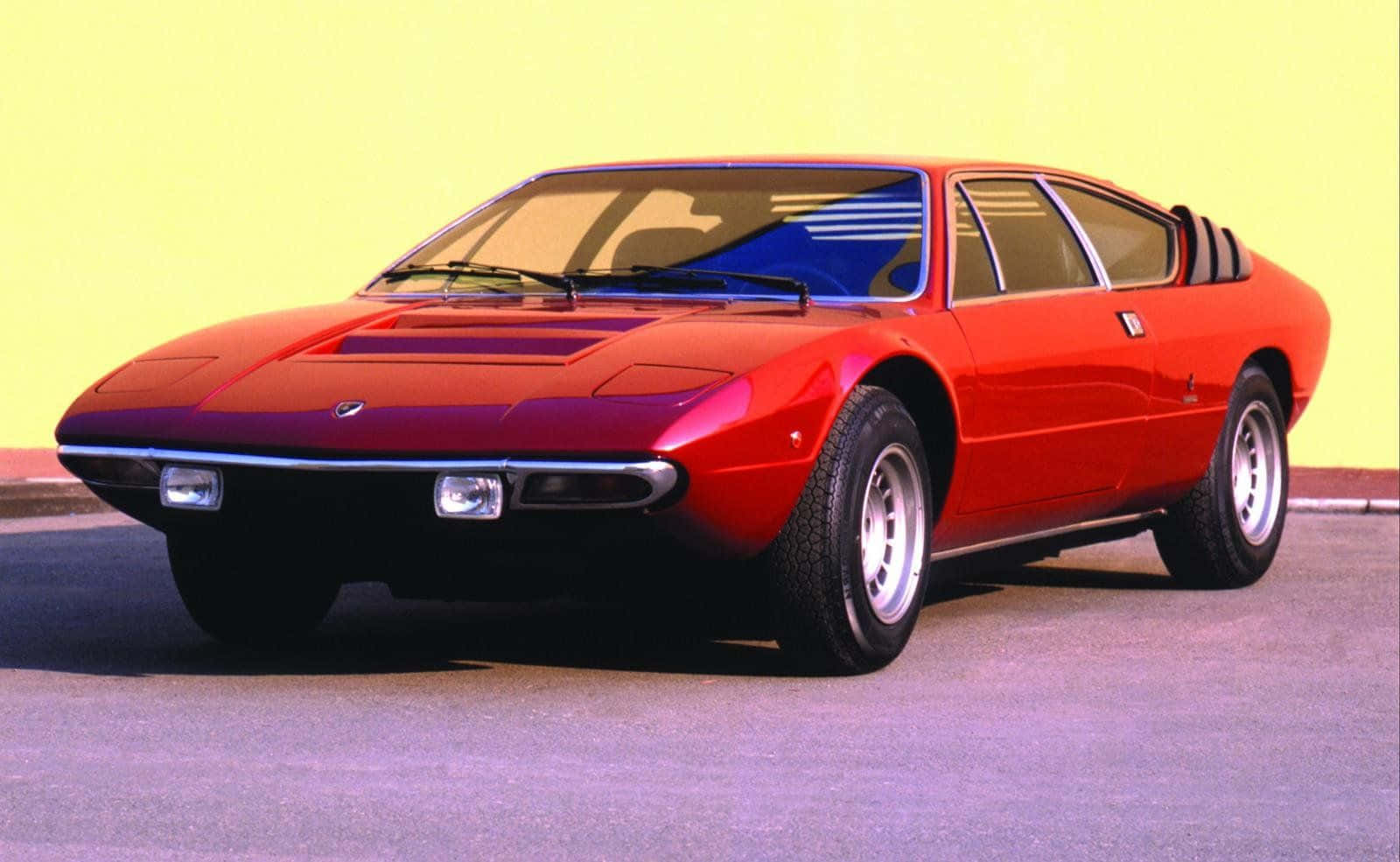 Lamborghini Urraco 桌布 壁紙 — 一輛充滿活力的紅色經典跑車，線條流暢，鍍鉻裝飾，在明亮的黃色工作室背景前展現風采。