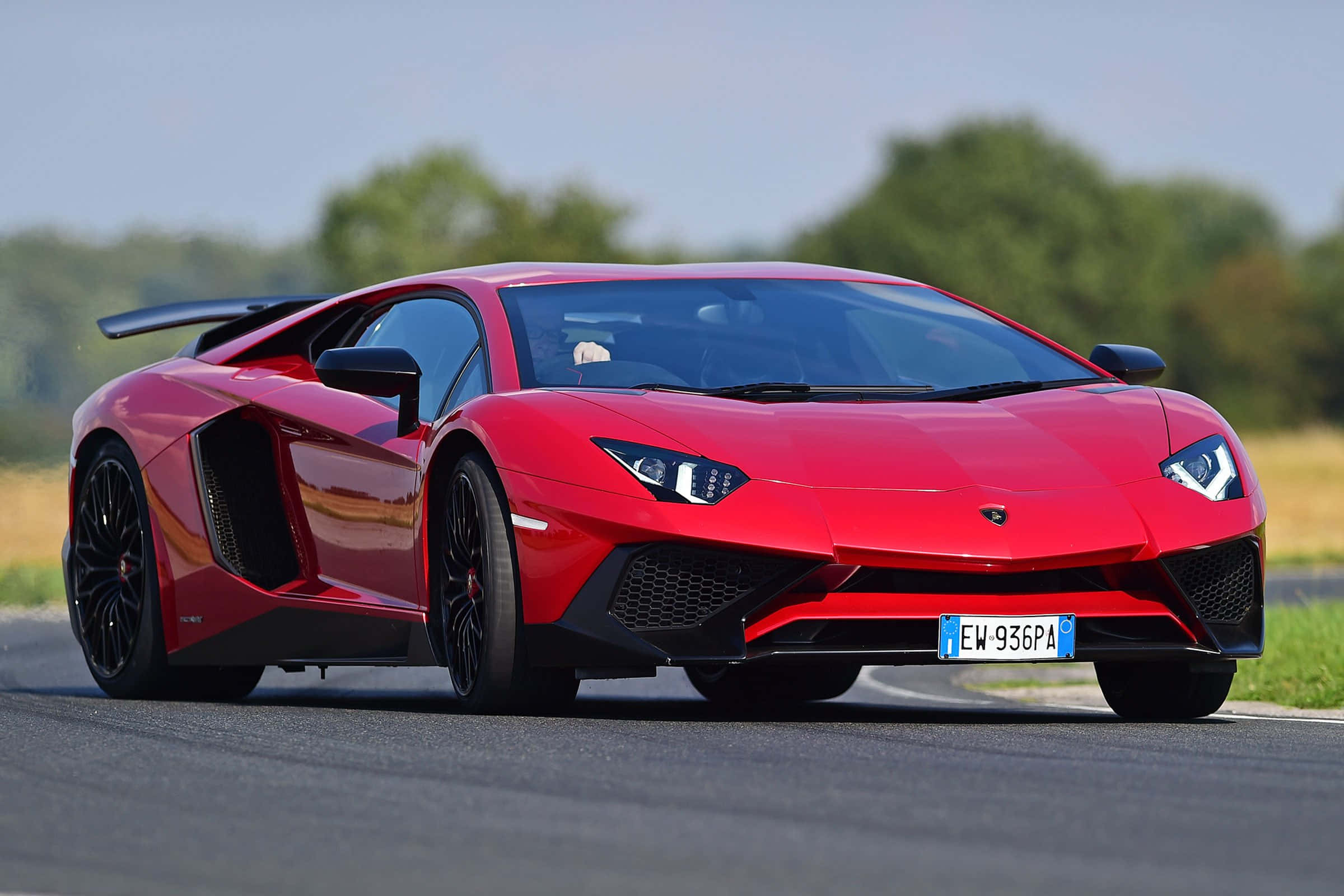 cars 桌布 壁紙 — 一輛充滿活力的紅色藍寶堅尼Aventador跑車，帶有黑色飾邊，在晴朗的天空下沿著一條兩旁綠樹成蔭的柏油道路奔馳。