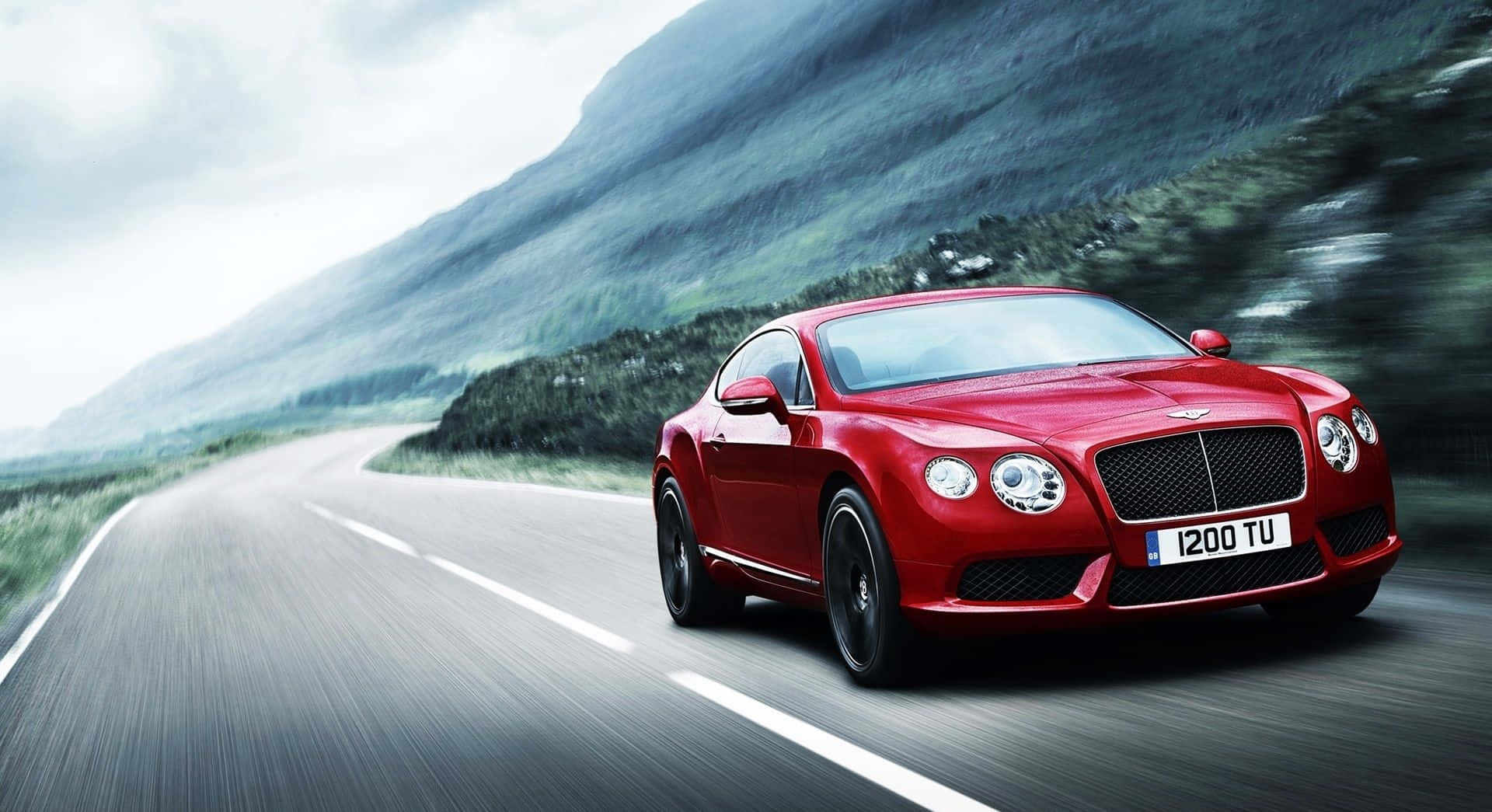 Best Bentley 桌布 壁紙 — 一輛充滿活力的紅色賓利 Continental GT 在蜿蜒的山路上疾馳，背景是模糊的綠意和霧氣繚繞的山峰。