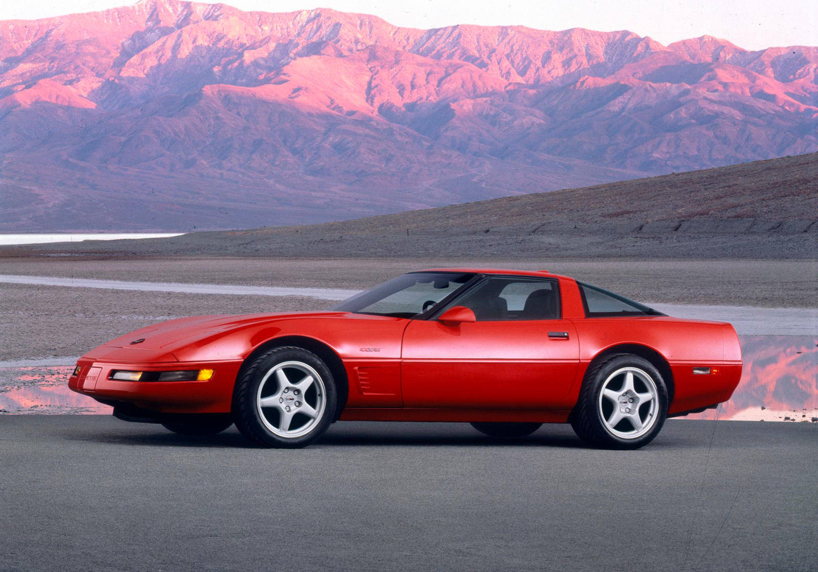 corvette 桌布 壁紙 — 一輛充滿活力的紅色跑車在夕陽下閃閃發光，背景是粉紅色的沙漠山脈。