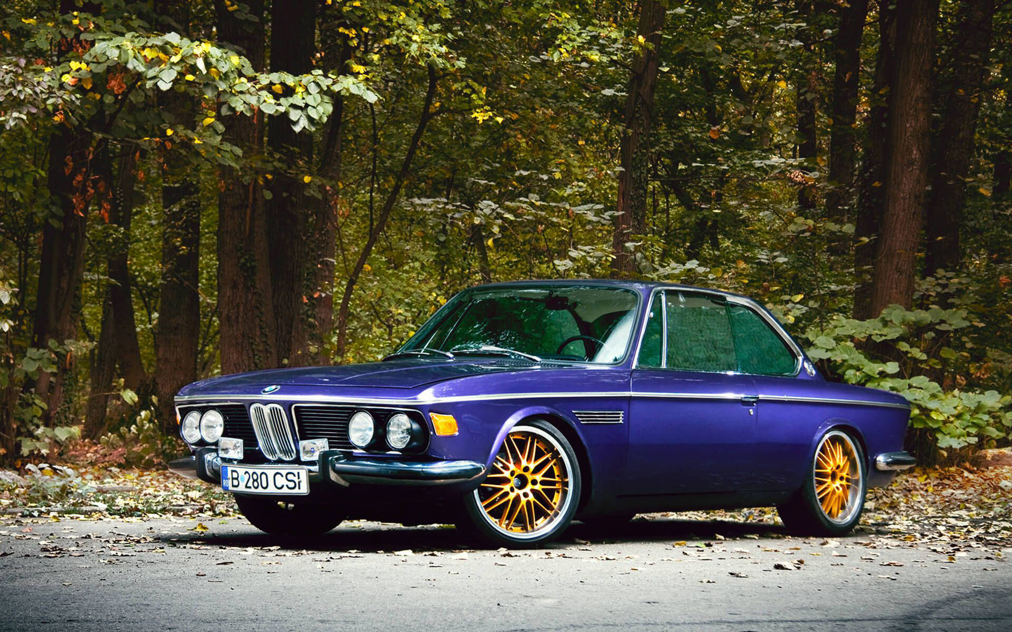 classic bmw 桌布 壁紙 — 一輛充滿活力的紫色經典BMW 280 CSI，配金色輪圈，停泊在秋樹下的森林道路上。
