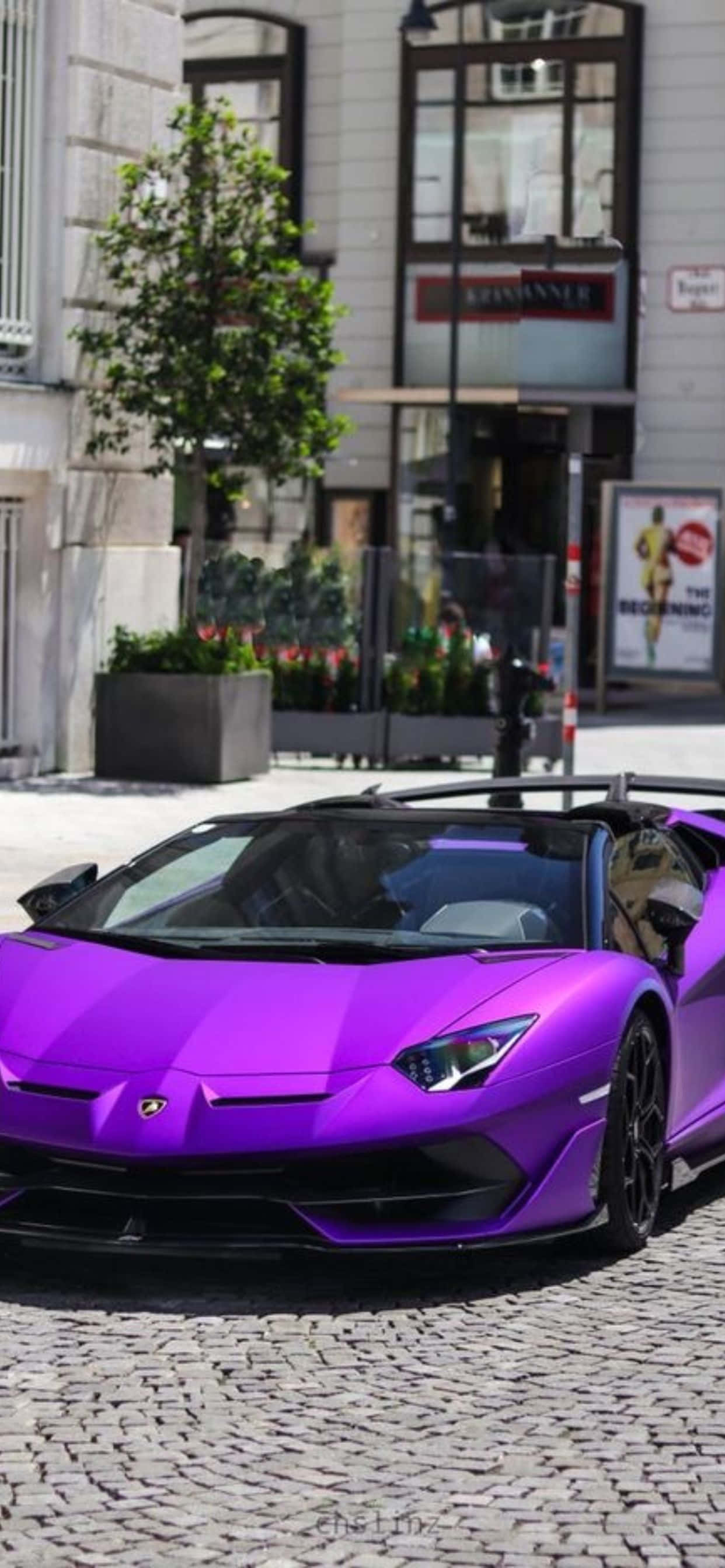 Lamborghini Phone 桌布 壁紙 — 一輛充滿活力的紫色藍寶堅尼 Aventador 敞篷跑車停泊在鵝卵石街道上，前方是一座帶有綠植的現代建築。