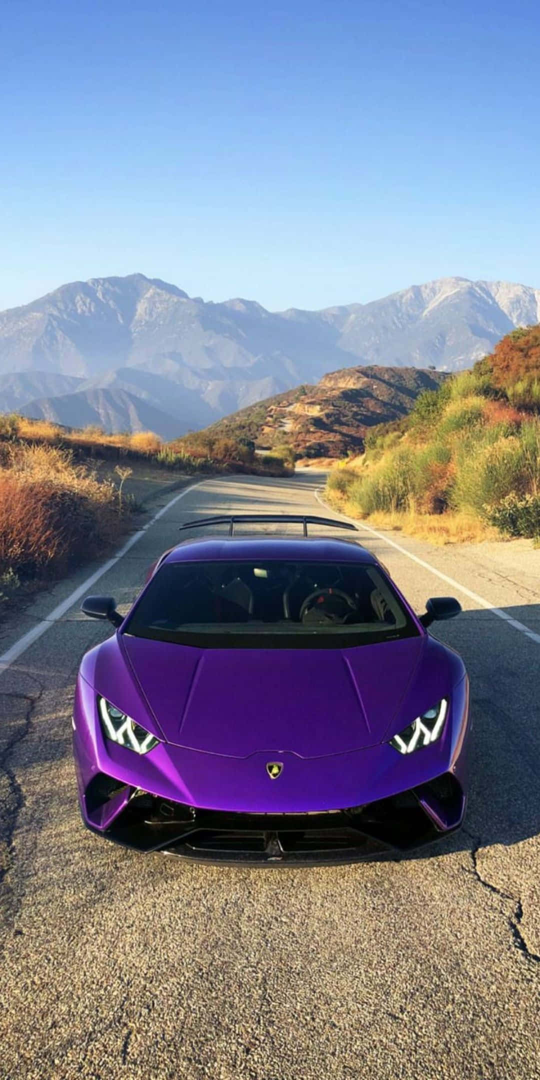Pixel 3 Lamborghini 桌布 壁紙 — 一輛充滿活力的紫色藍寶堅尼Huracán停泊在蜿蜒的山路上，背景是晴朗的藍天和遠山。