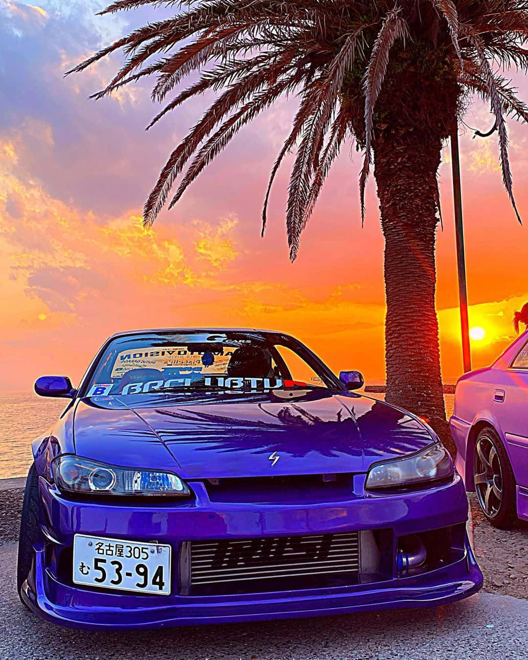 JDM iPhone 桌布 壁紙 — 一輛充滿活力的紫色跑車停放在棕櫚樹下，夕陽映照出火熱的橙色和粉紅色天空。
