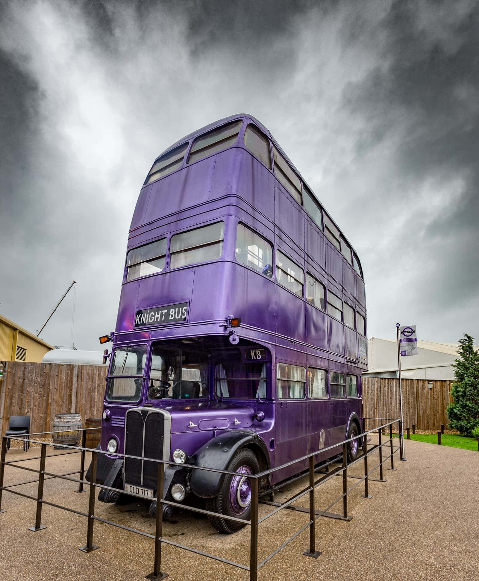 The Hogwarts Knight Bus Stop 桌布 壁紙 — 一輛充滿活力的紫色雙層巴士，標有「Knight Bus」，矗立在戲劇性的多雲天空下，被金屬圍欄包圍。
