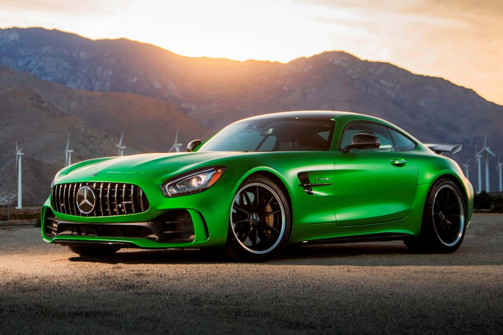 汽車 桌布 壁紙 — 一輛充滿活力的綠色 Mercedes-AMG GT 跑車在金色夕陽下閃耀，背景是山脈和風力發電機。