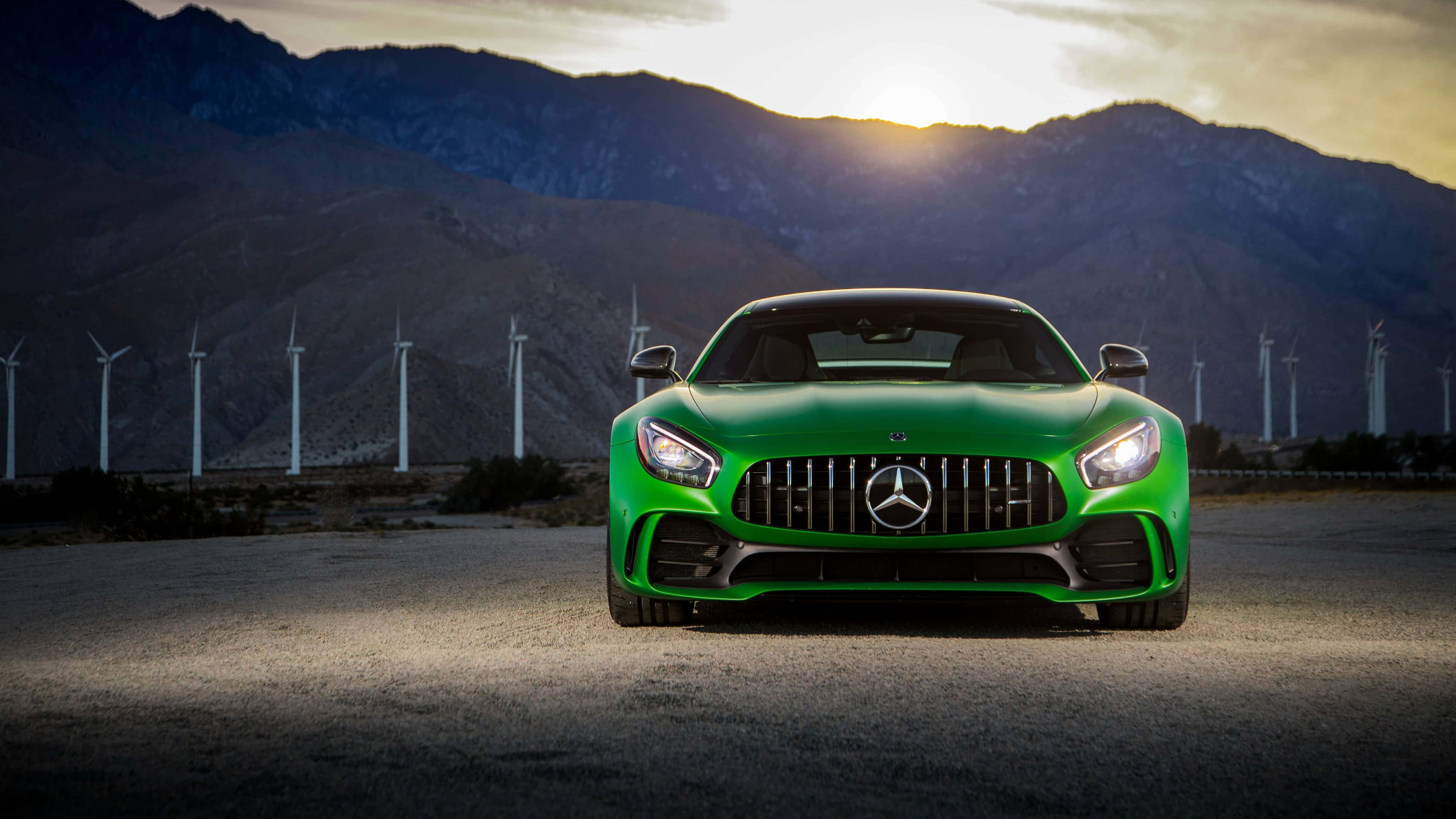 Mercedes 桌布 壁紙 — 一輛充滿活力的綠色 Mercedes-AMG GT 跑車在沙漠公路上向前行駛，背景是風力發電機和山脈，金色夕陽映照。