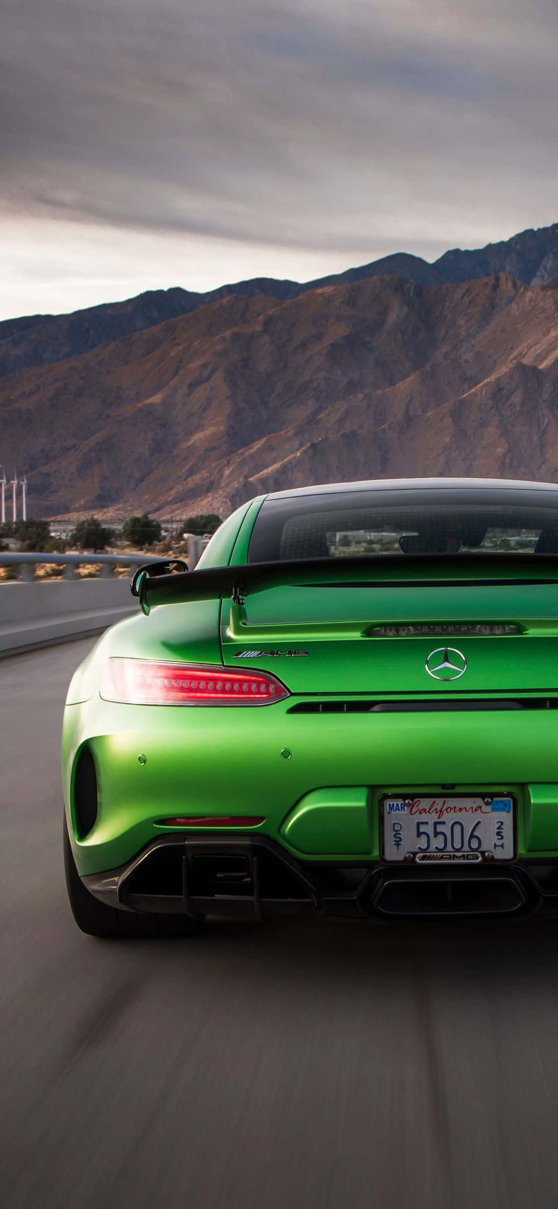 山景 桌布 壁紙 — 一輛充滿活力的綠色 Mercedes-AMG GT R 跑車在沙漠公路上疾馳，背景是崎嶇的山脈和陰雲密布的天空。