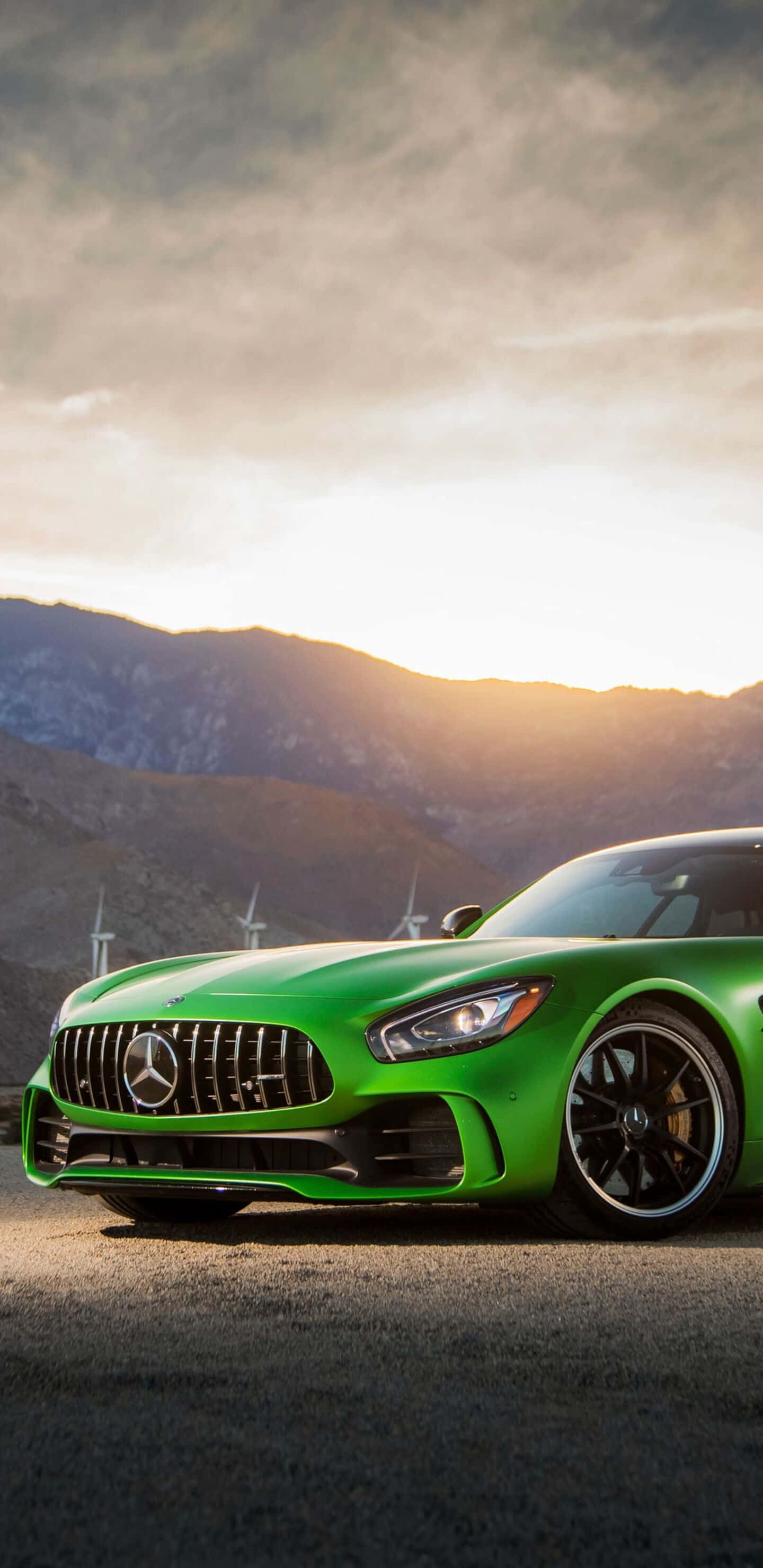 Car Images HD 桌布 壁紙 — 一輛充滿活力的綠色 Mercedes-AMG GT 跑車停泊在瀝青路上，背景有風力發電機和山脈，天空呈現金色日落。