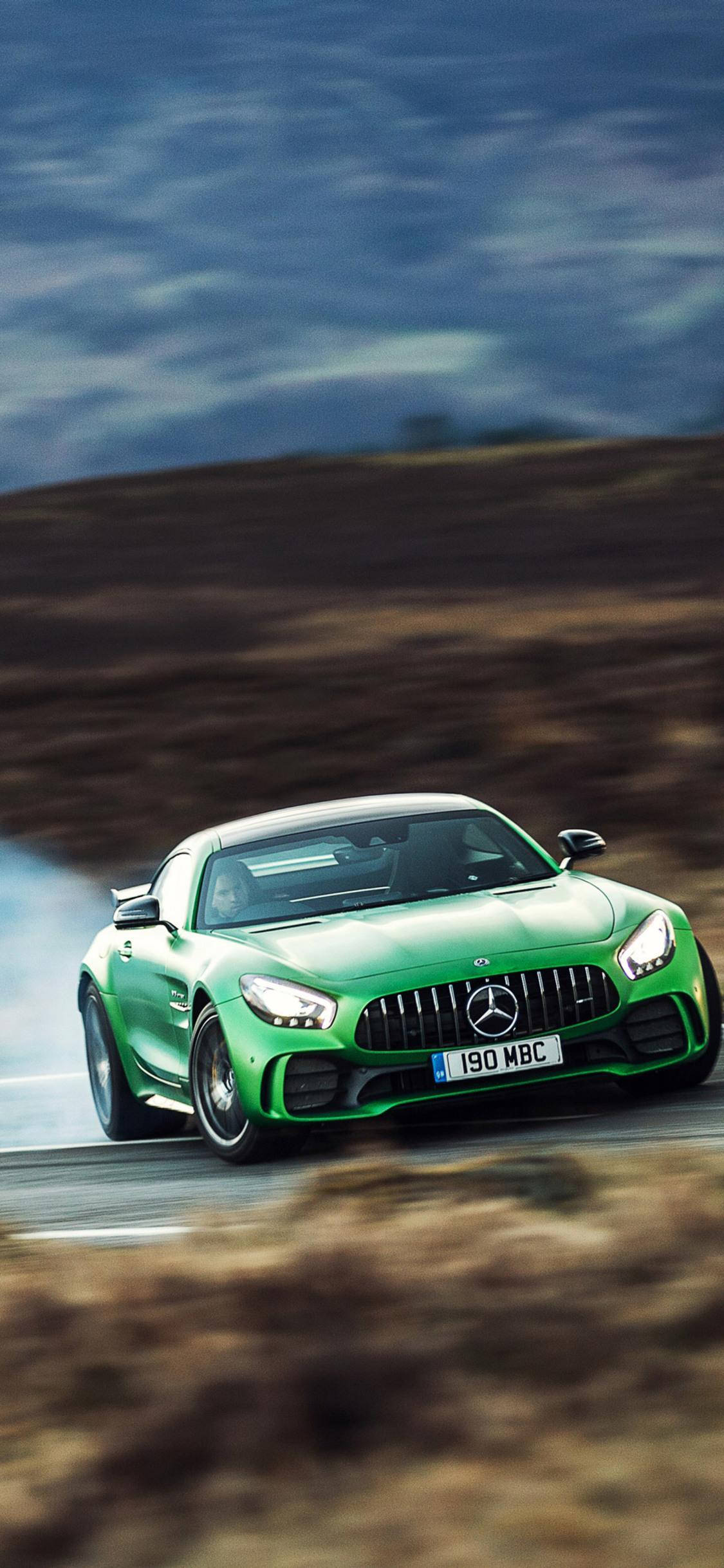 amg iPhone 桌布 壁紙 — 一輛充滿活力的綠色 Mercedes-AMG GT R 跑車，在崎嶇景觀中的蜿蜒道路上，於陰鬱的天空下強力飄移。