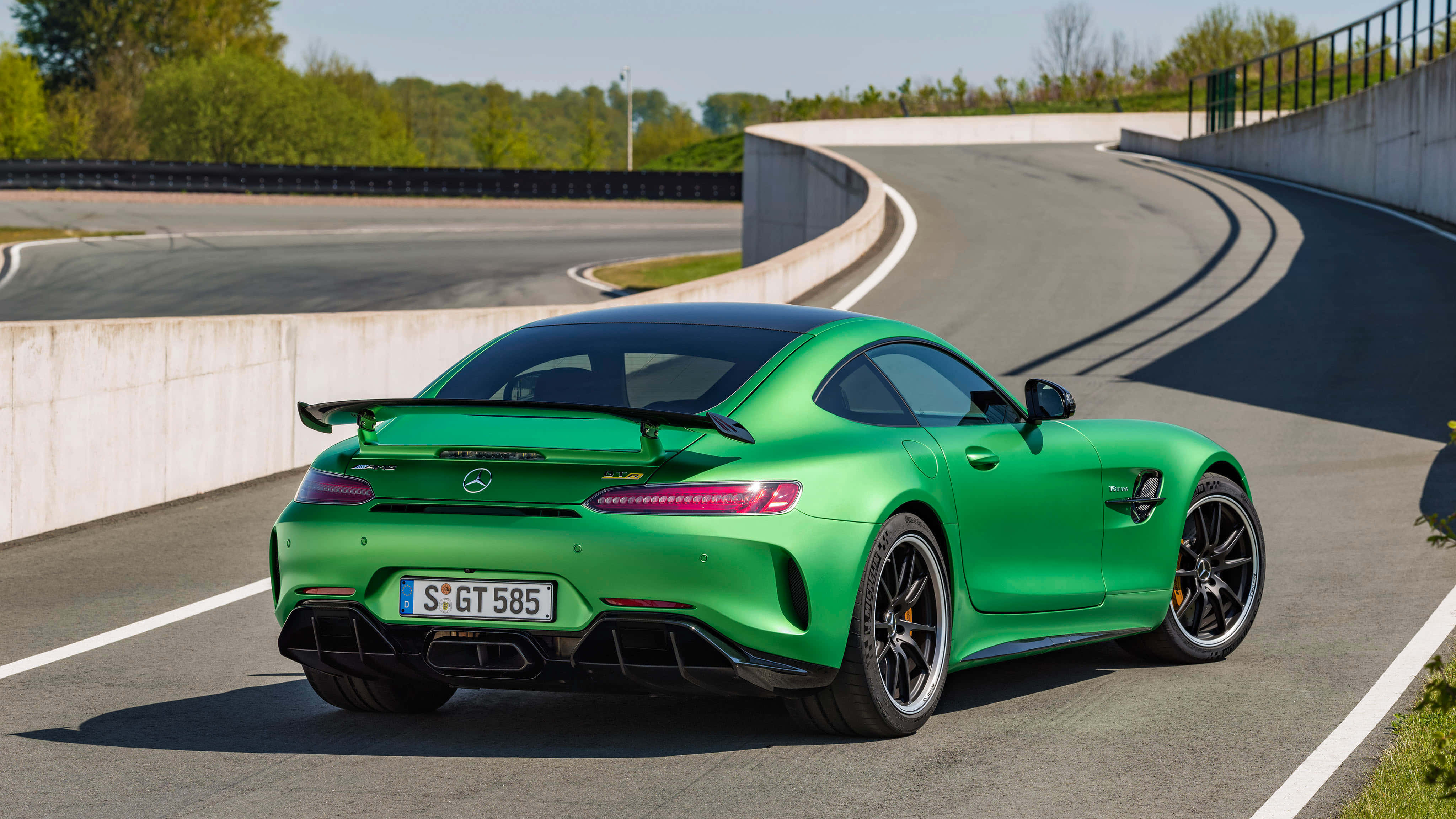 Road 桌布 壁紙 — 一輛充滿活力的綠色 Mercedes-AMG GT R 跑車在晴朗天空下，沿著由混凝土護欄和綠樹環繞的彎曲瀝青賽道行駛。