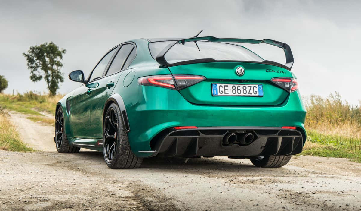 愛快羅密歐 桌布 壁紙 — 一輛充滿活力的綠色Alfa Romeo Giulia GTA跑車，帶有大型後擾流板，停泊在泥路上，周圍是草地和樹木，天空多雲。