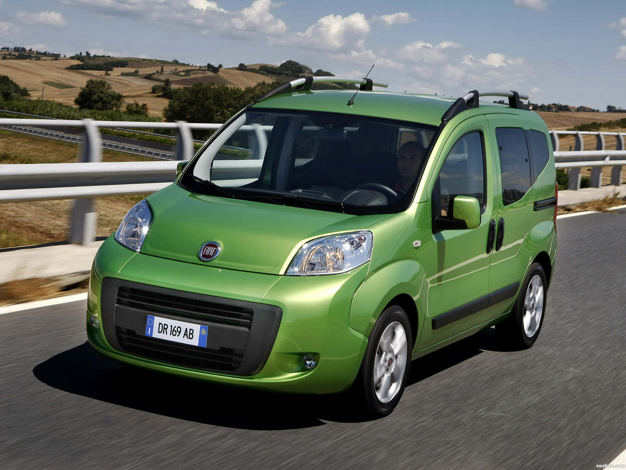 Fiat Fiorino 桌布 壁紙 — 一輛充滿活力的綠色Fiat Panda廂型車在陽光明媚的鄉村高速公路上行駛，背景是起伏的丘陵和散落的樹木。
