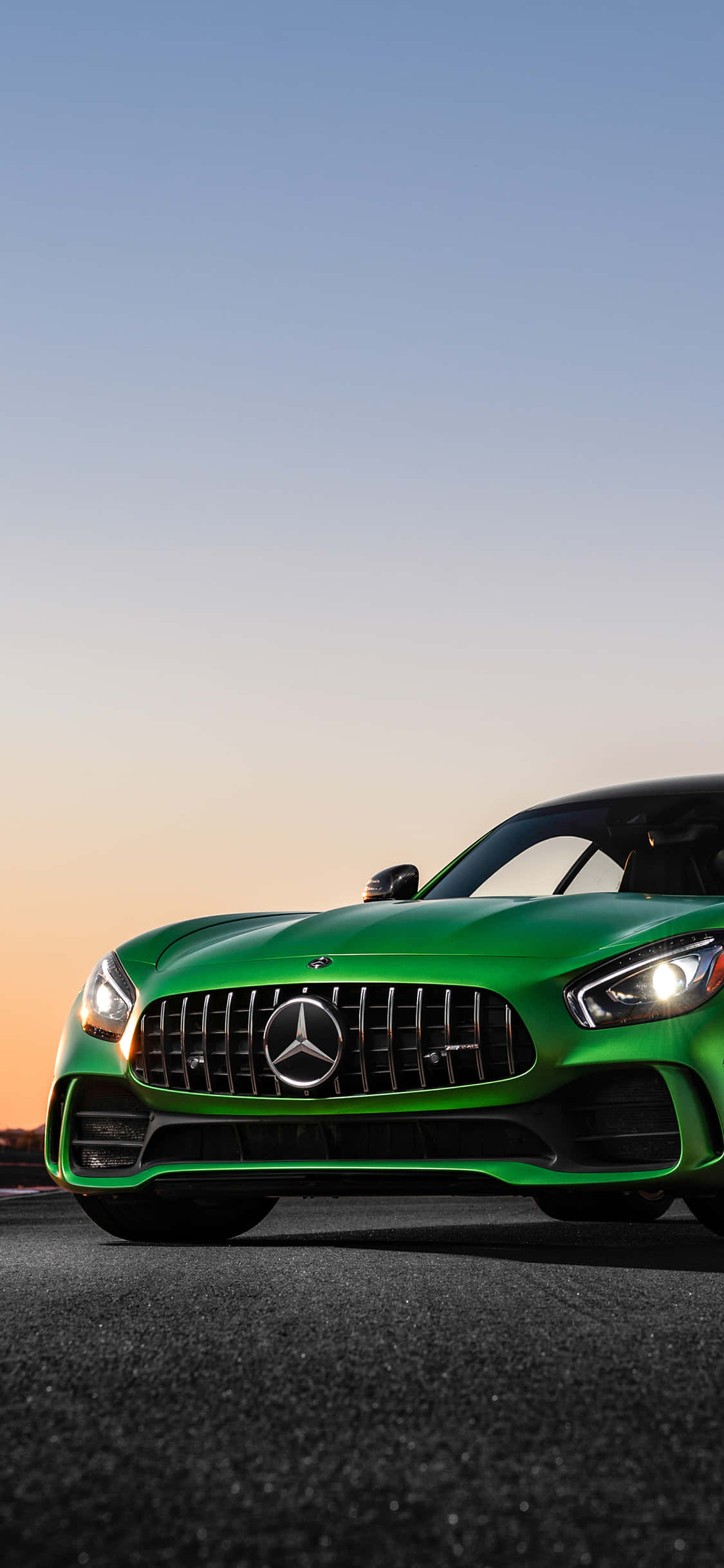iPhone X Amg Gt-r 桌布 壁紙 — 一輛充滿活力的綠色Mercedes-AMG GT跑車在柔和的日落天空下於瀝青路上閃閃發光。