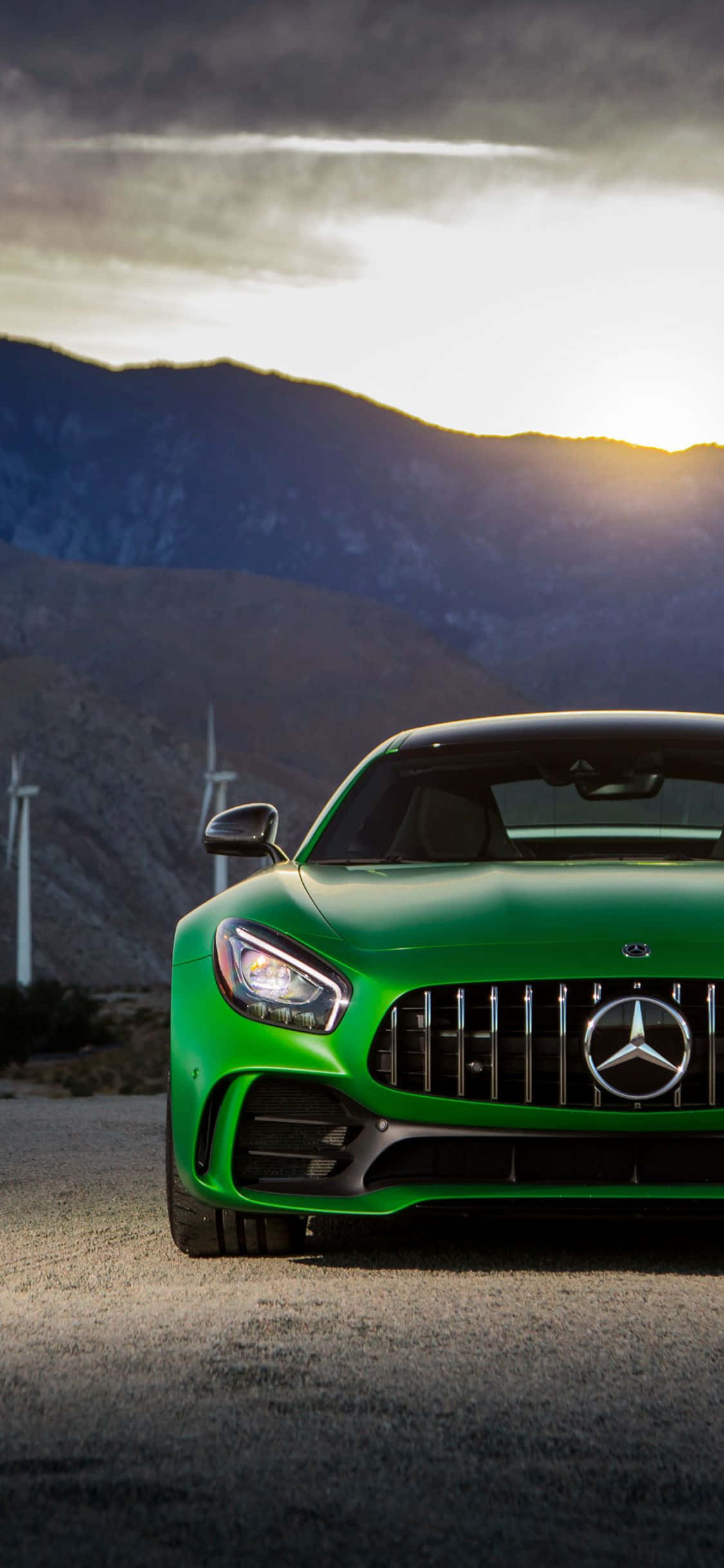 Mercedes 桌布 壁紙 — 一輛充滿活力的綠色Mercedes-AMG GT跑車停泊在碎石路上，背景是風力發電機和山脈，天空呈現戲劇性的日落景色。