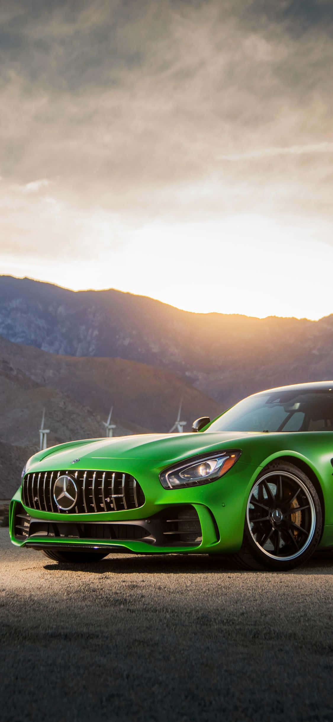 Mercedes 桌布 壁紙 — 一輛充滿活力的綠色Mercedes-AMG GT跑車停在瀝青路上，背景有風力發電機和山脈，天空呈現金色日落。