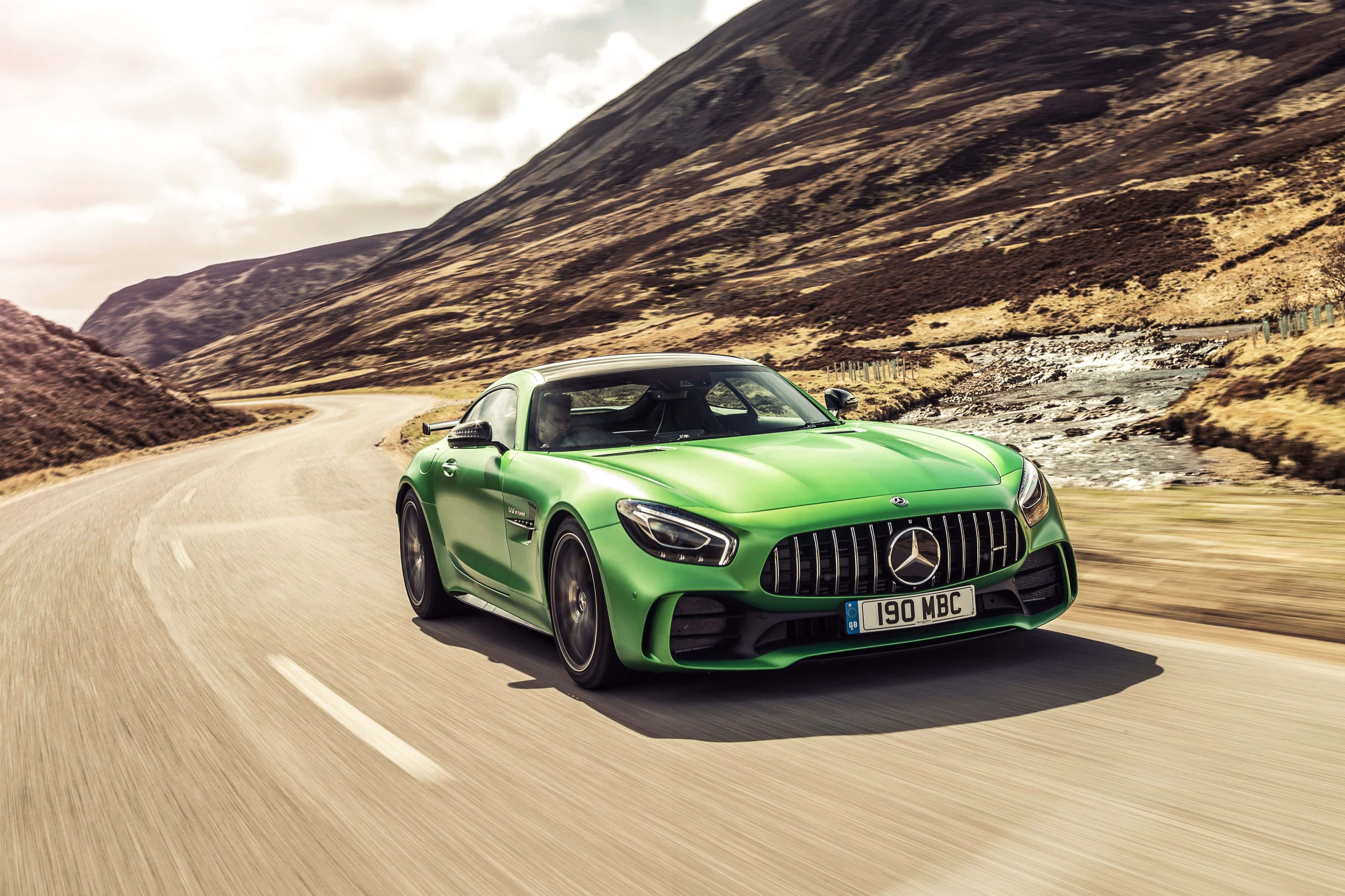 Mercedes 桌布 壁紙 — 一輛充滿活力的綠色Mercedes-AMG GT跑車沿著蜿蜒的山路飛馳，背景是崎嶇的山丘和一條河流。